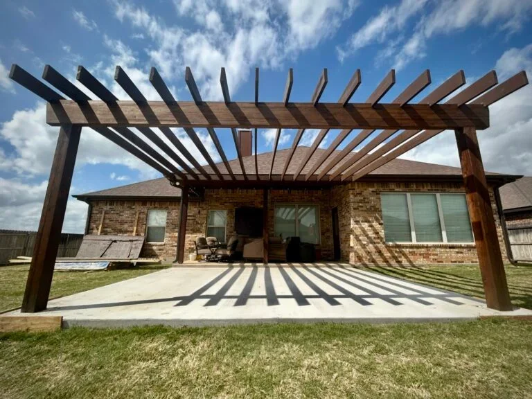 Backyard patio with a wooden pergola casting shadows on the concrete floor, attached to a brick house under a partly cloudy sky.