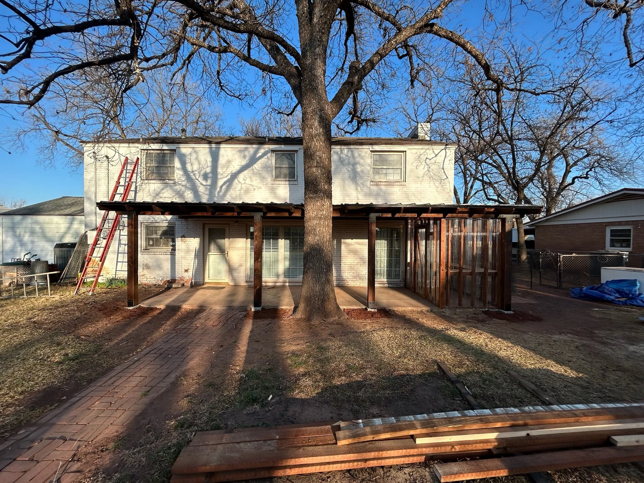 The back view of a two-story house with a large tree in front, a brick-paved pathway, a ladder leaning against the house, and construction materials in the yard, indicating ongoing renovation or construction work.
