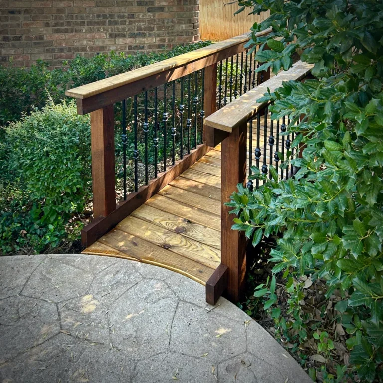 A small wooden bridge with black metal railing in a garden, surrounded by green bushes and plants.