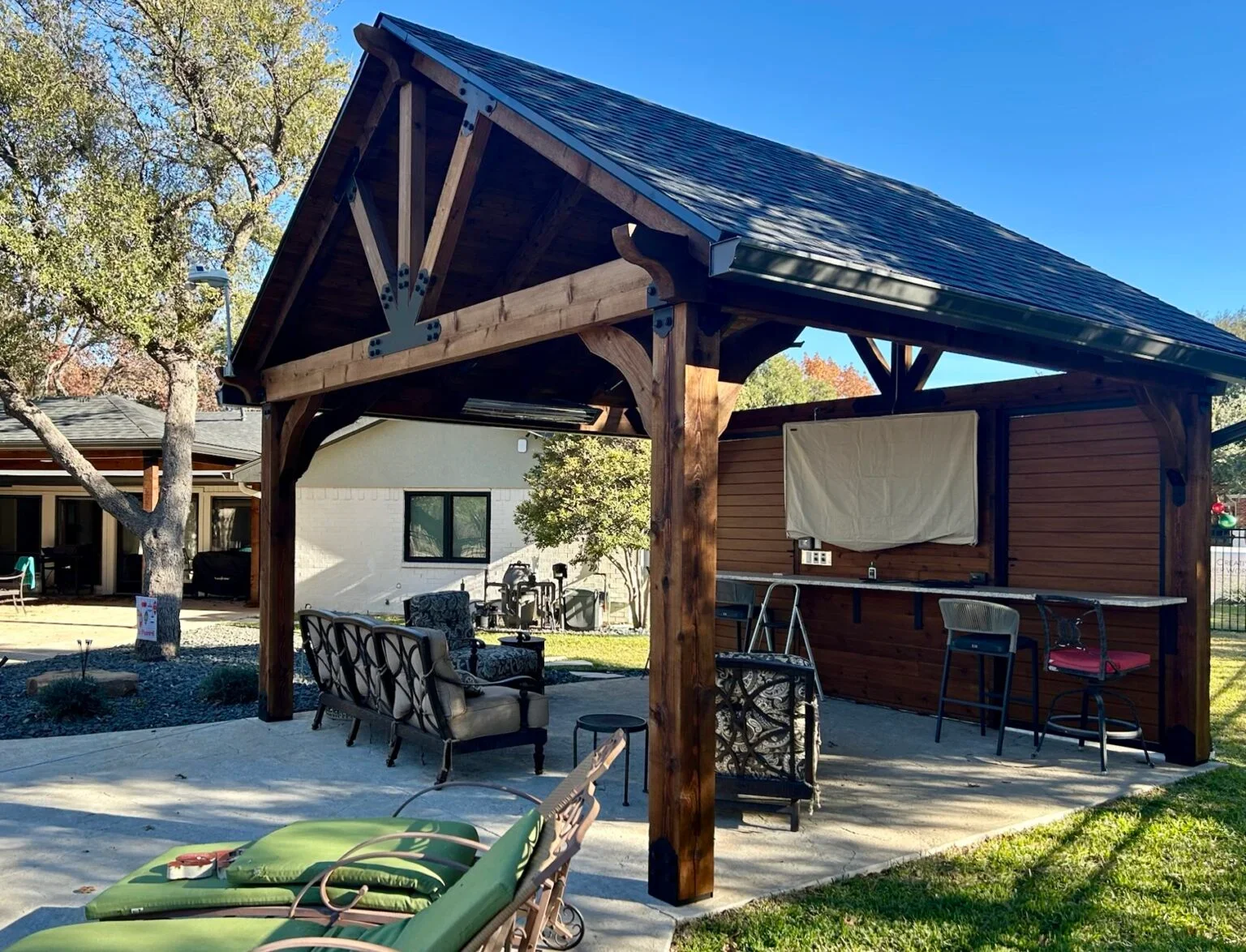 An outdoor covered patio with wooden beams and a blue shingled roof. The patio has seating including chairs and a loveseat, a bar area with chairs, and surrounding trees and houses in the background.
