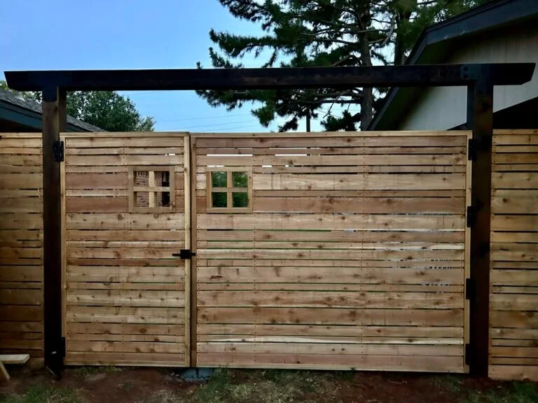 A wooden backyard fence with an archway and a gate, featuring two small window sections, set against trees and a house in the background.