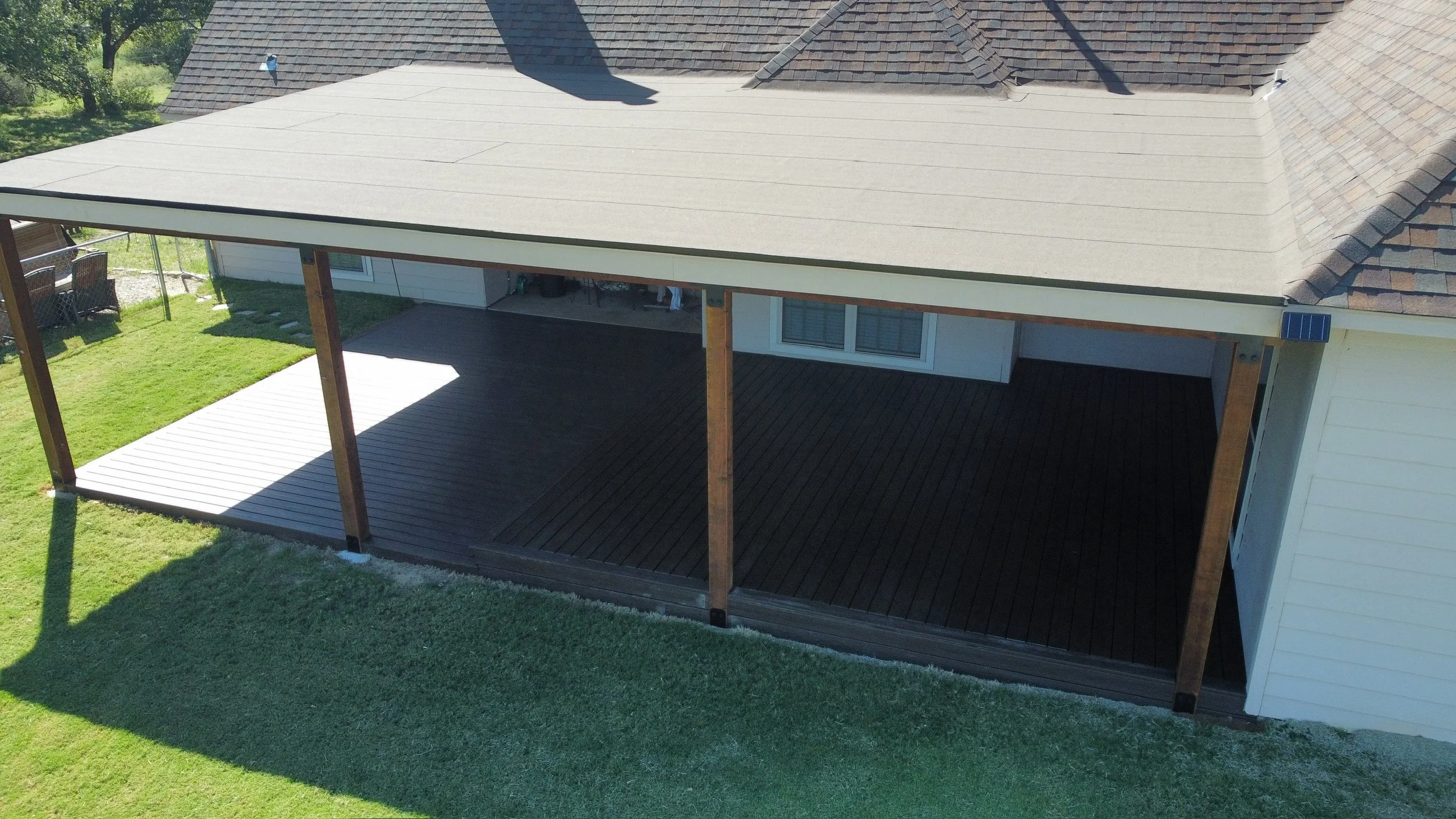 A house's backyard with a covered deck and wooden flooring, surrounded by green grass and trees.