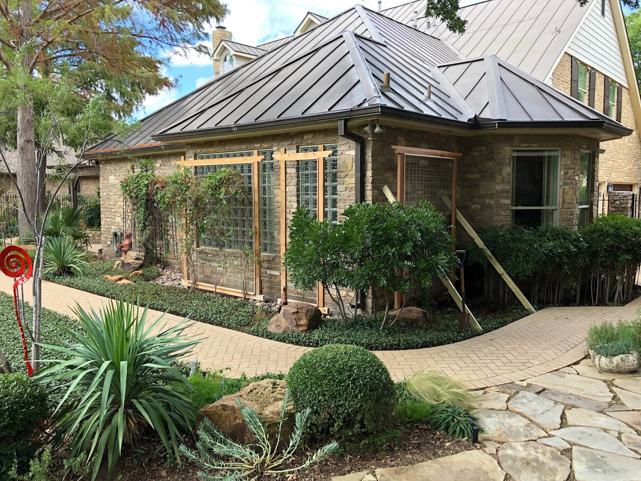 A backyard garden area with a brick house featuring a metal roof, surrounded by green plants, trees, and a paved pathway.