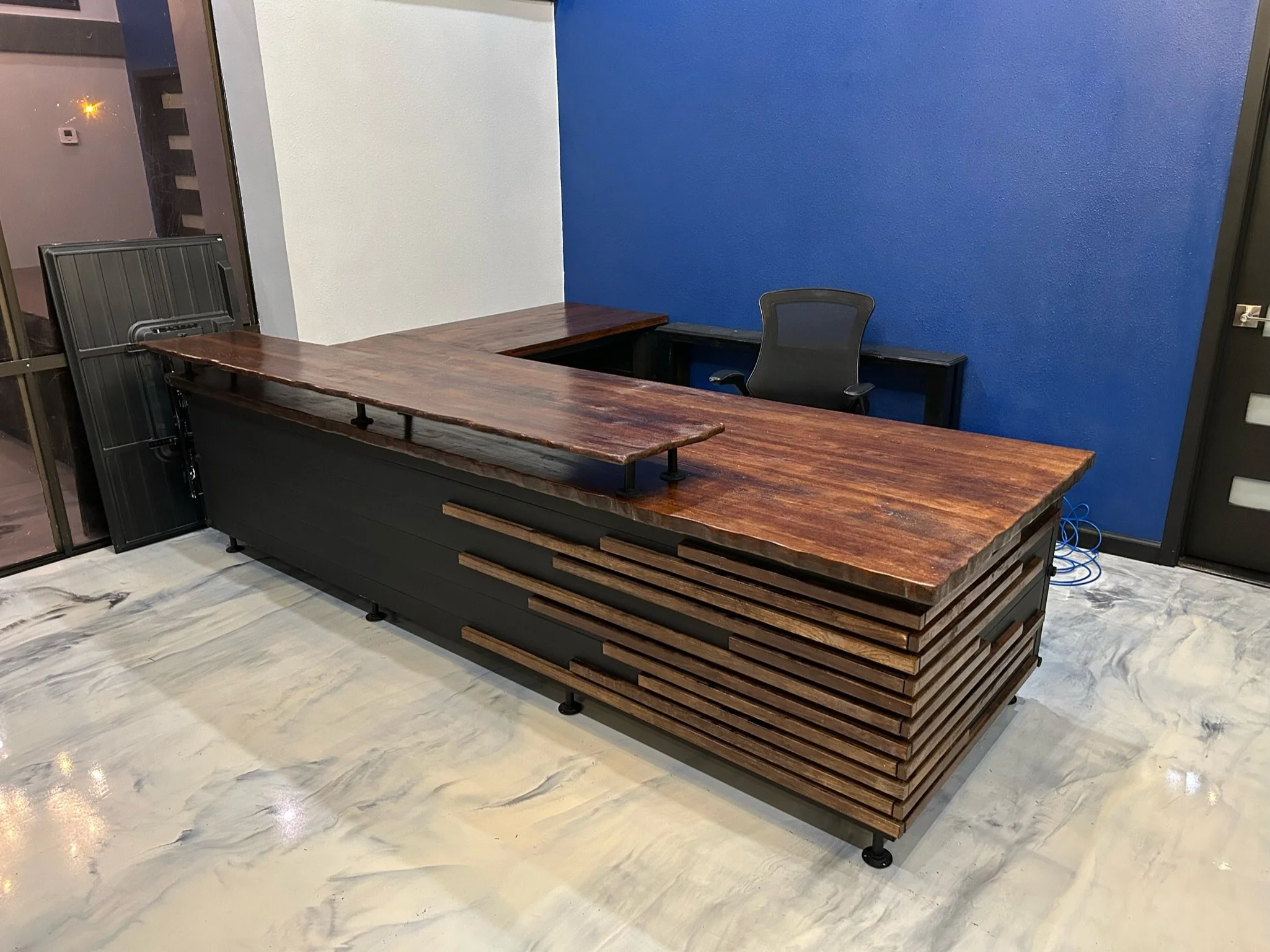 An empty office space with a desk made of wood and metal, a black office chair, and a blue accent wall. There is a doorway with a black door frame and some electrical cords on the floor.