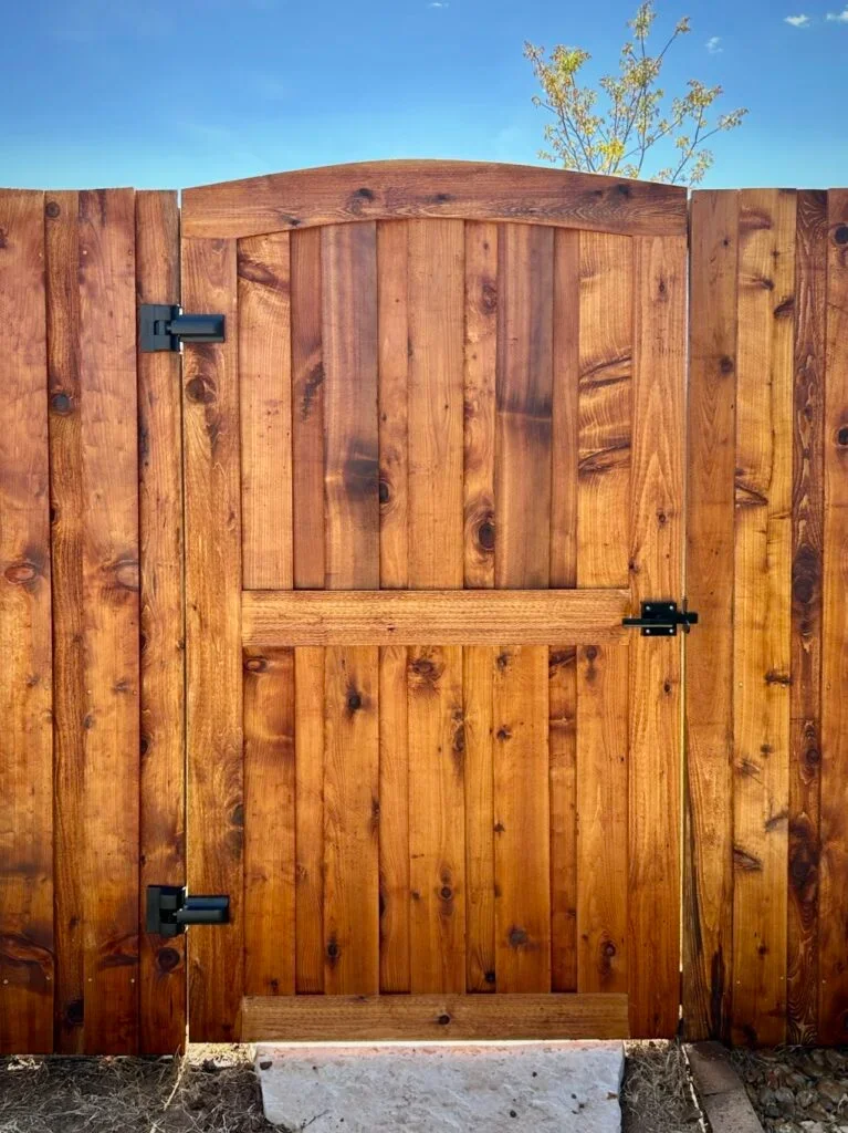 A wooden gate with black hinges and latch, part of a wooden fence, under a blue sky with a tree in the background.