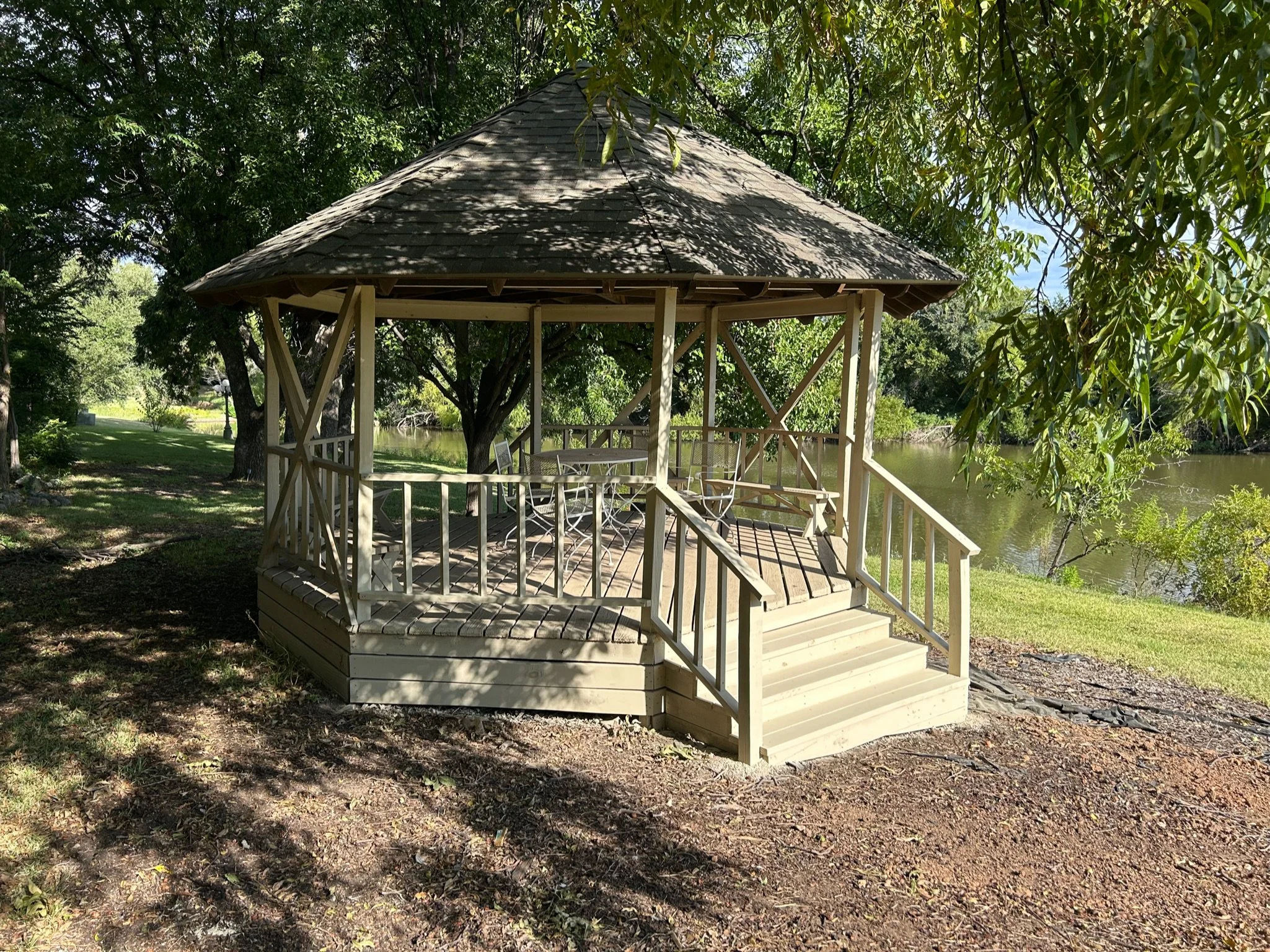 Wooden gazebo by a river surrounded by green trees.