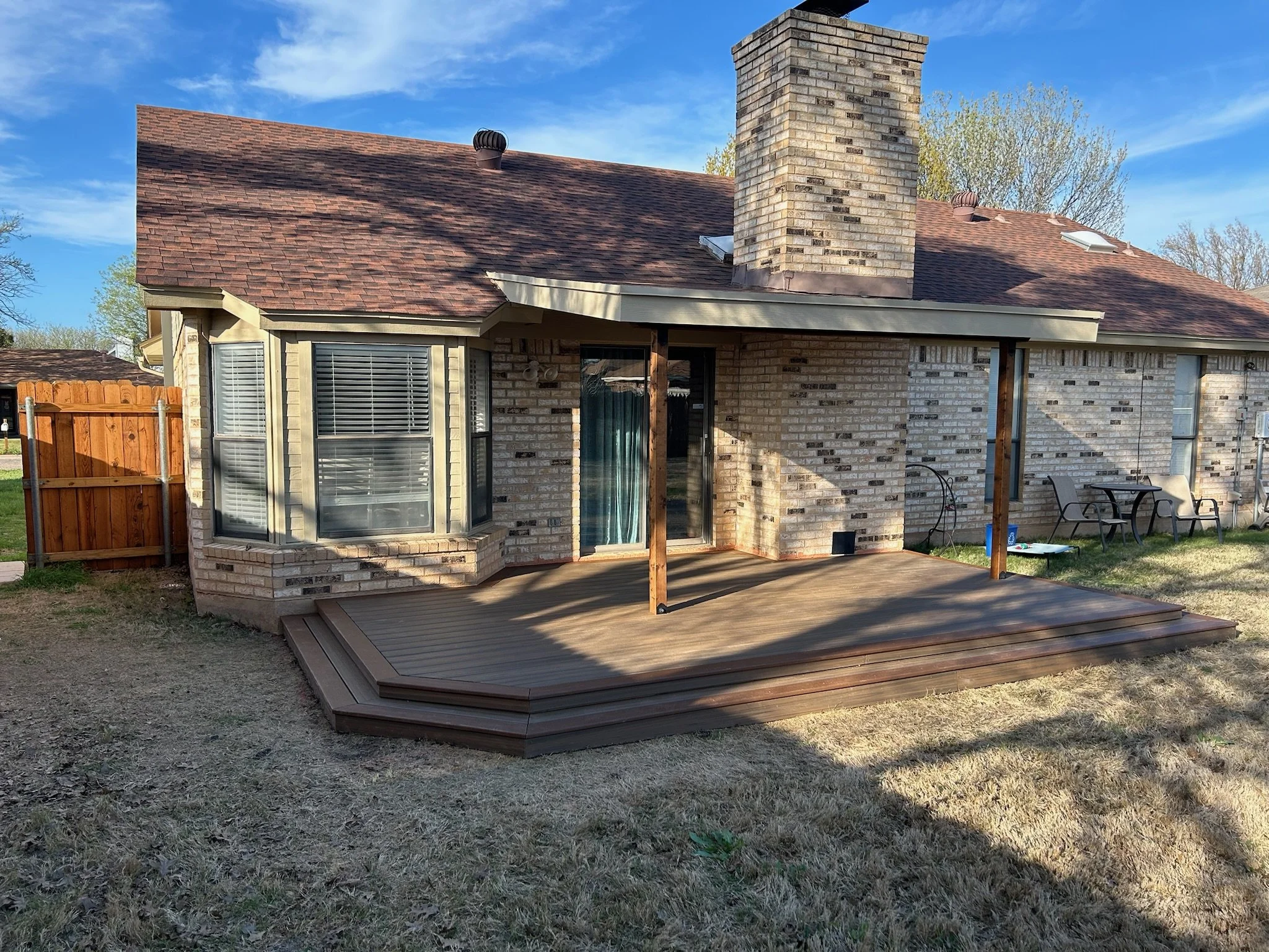 Backyard patio with a wooden deck, patio chairs, a brick house with large windows, a chimney, and a fence. There are trees and a bright blue sky with some clouds.