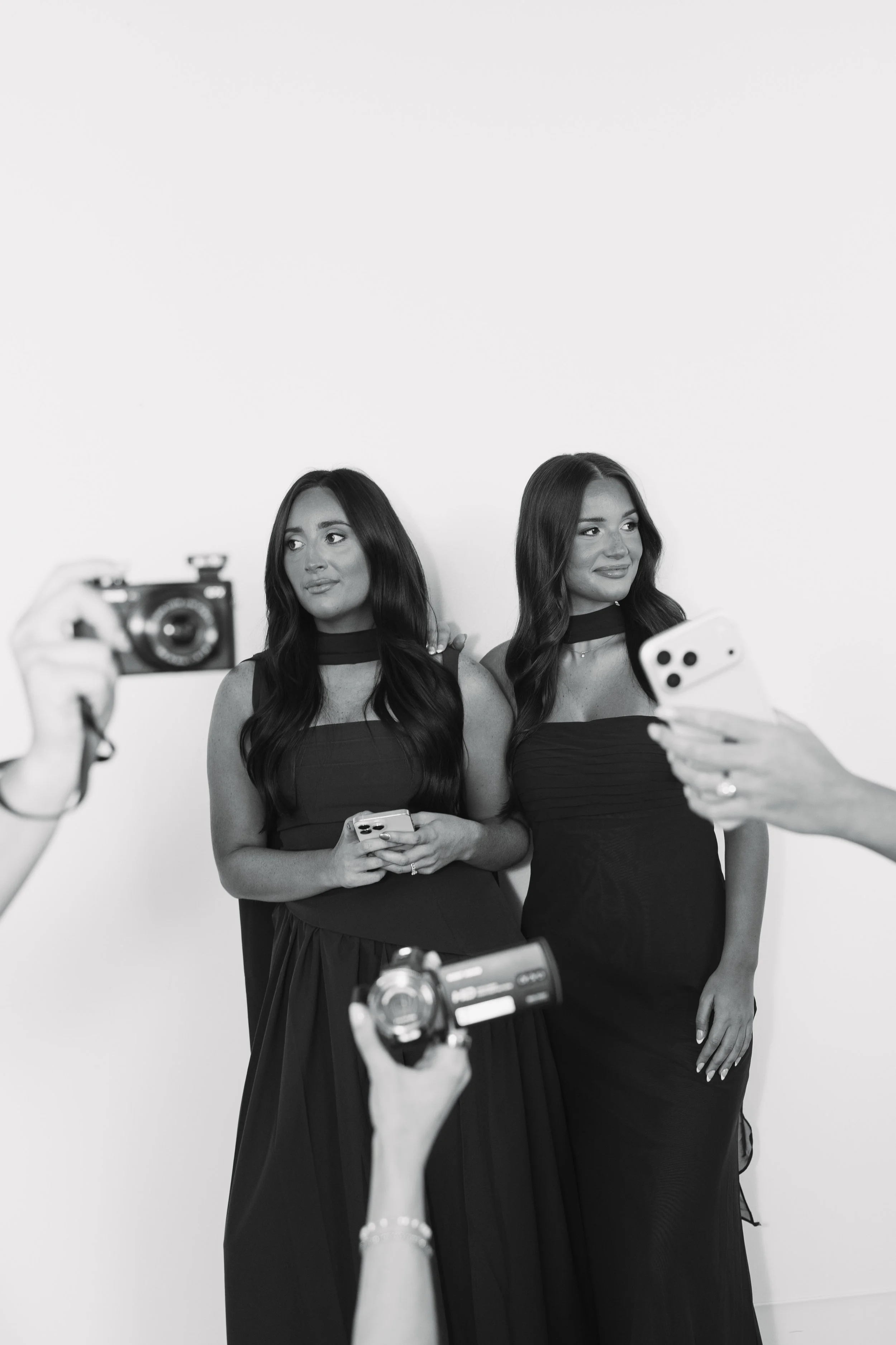 Two women in black dresses posing for photos in front of a plain background, one holding a phone and the other holding a smartphone, while others take pictures of them.