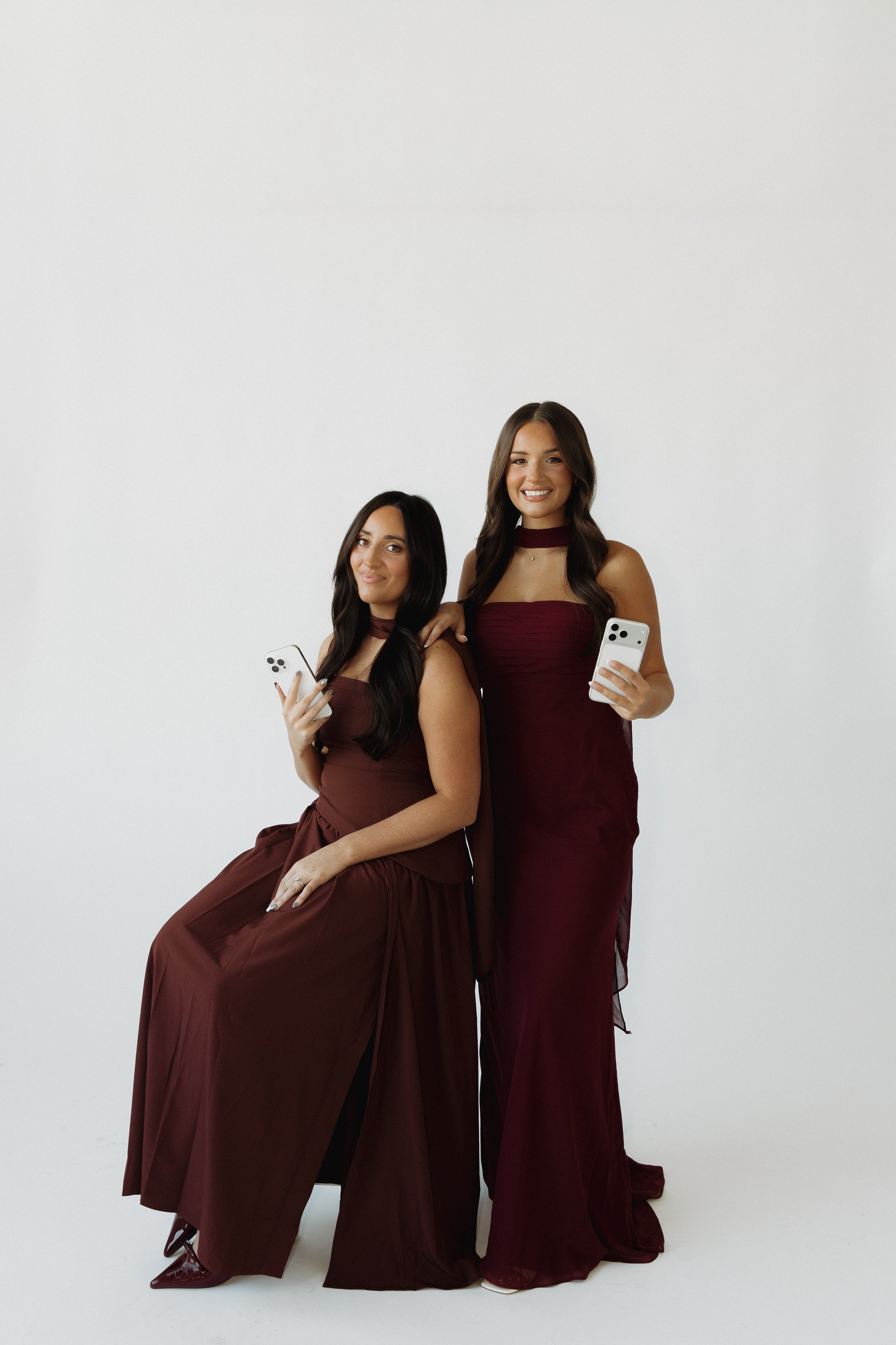 Two women in matching wine-colored dresses holding smartphones, standing against a plain white background.