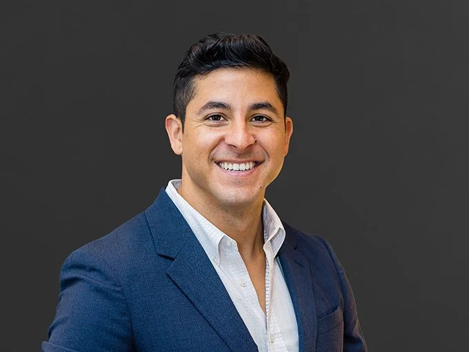Portrait of a man in a blue suit smiling against a dark gray background.