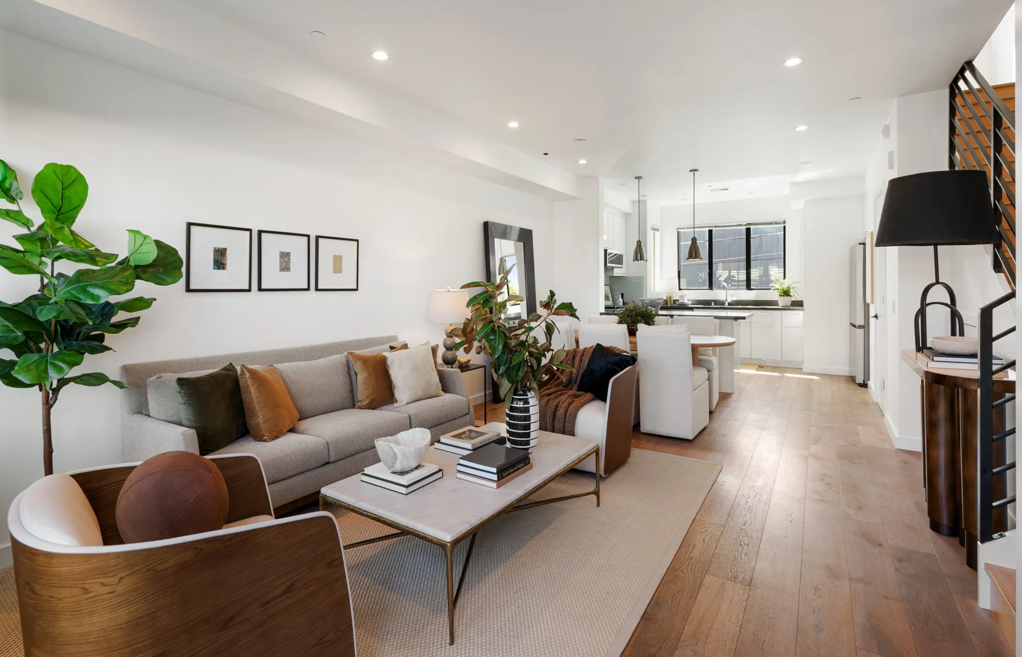 Interior of a modern living room with a beige sofa, white walls, framed artwork, green plants, wooden flooring, and a view of the kitchen and dining area in the background.