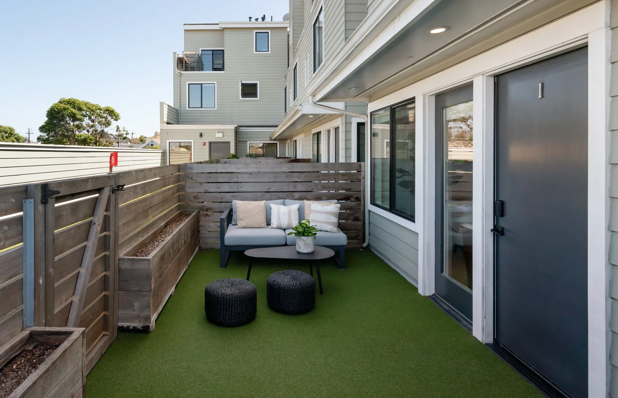 Small balcony with artificial grass flooring, black outdoor furniture including a sofa with pillows, a small round table with a potted plant, and two black ottomans. Wooden privacy fence on one side, gray side door, sliding glass doors on the buildin