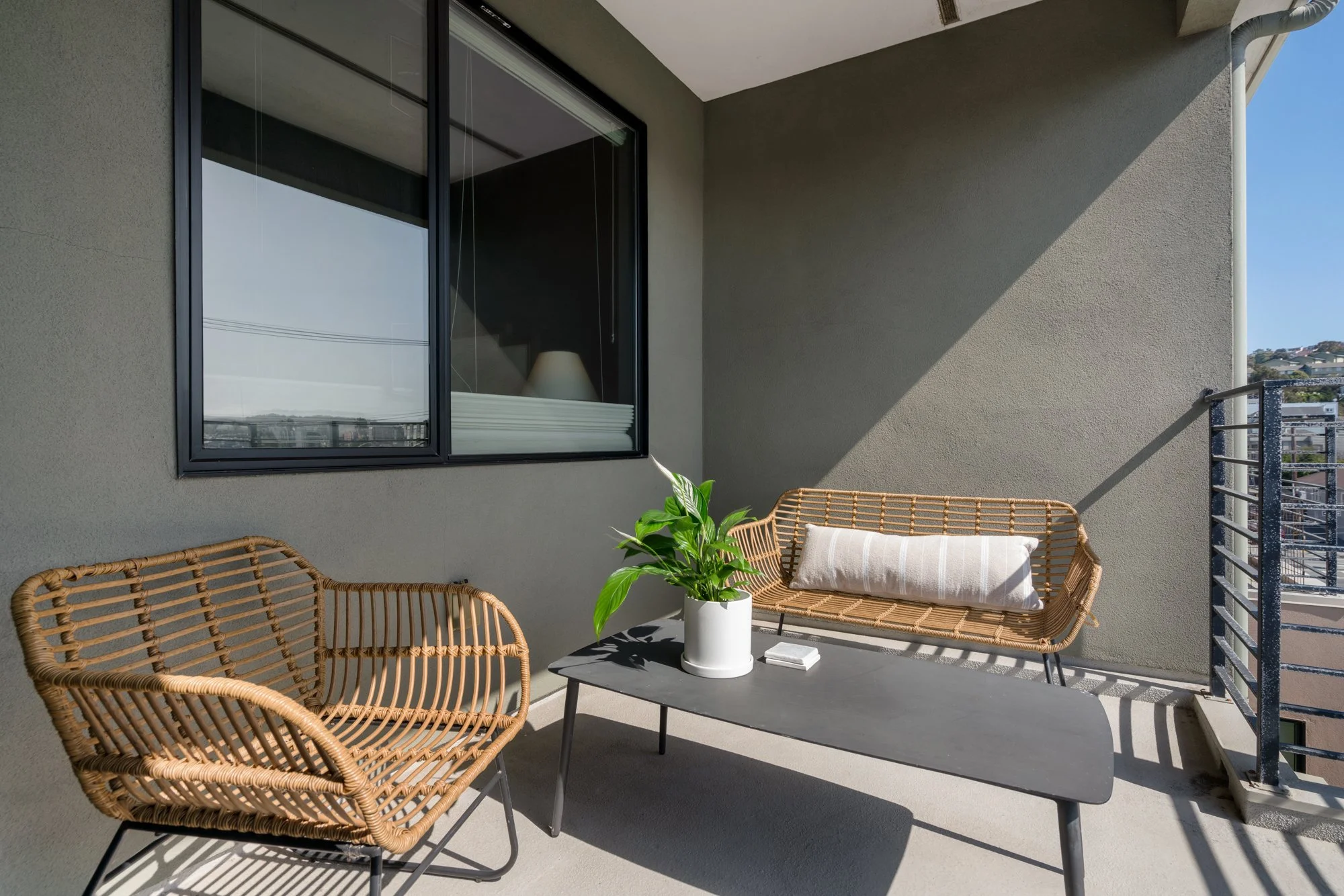 Balcony with wicker chairs, a coffee table with a potted plant and sunglasses, and a window showing a reflection of the sky and horizon.