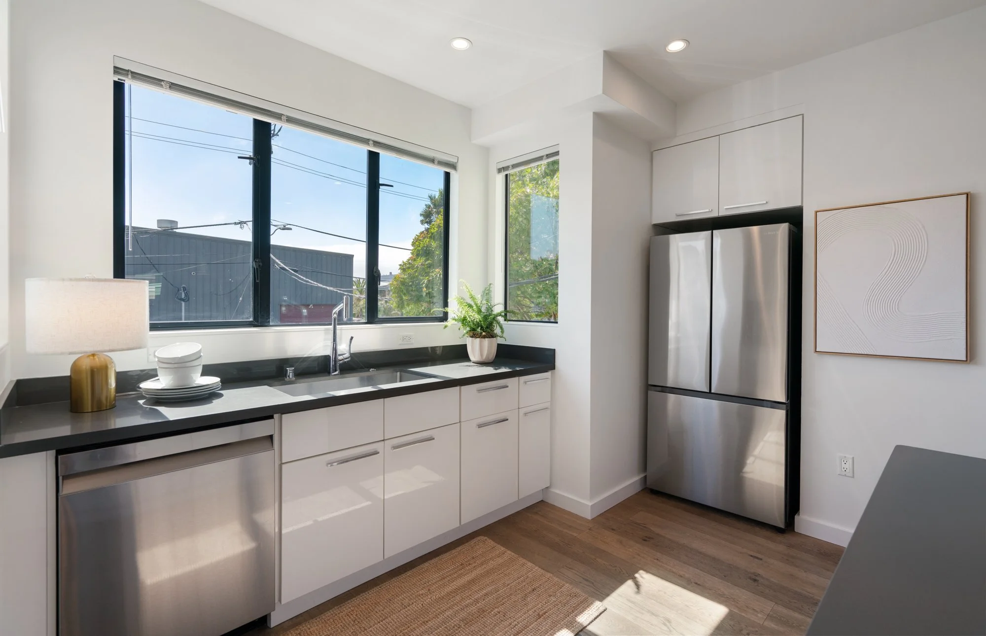 Modern kitchen with large window, white cabinets, stainless steel refrigerator, black countertop, potted plant, table lamp, and abstract wall art