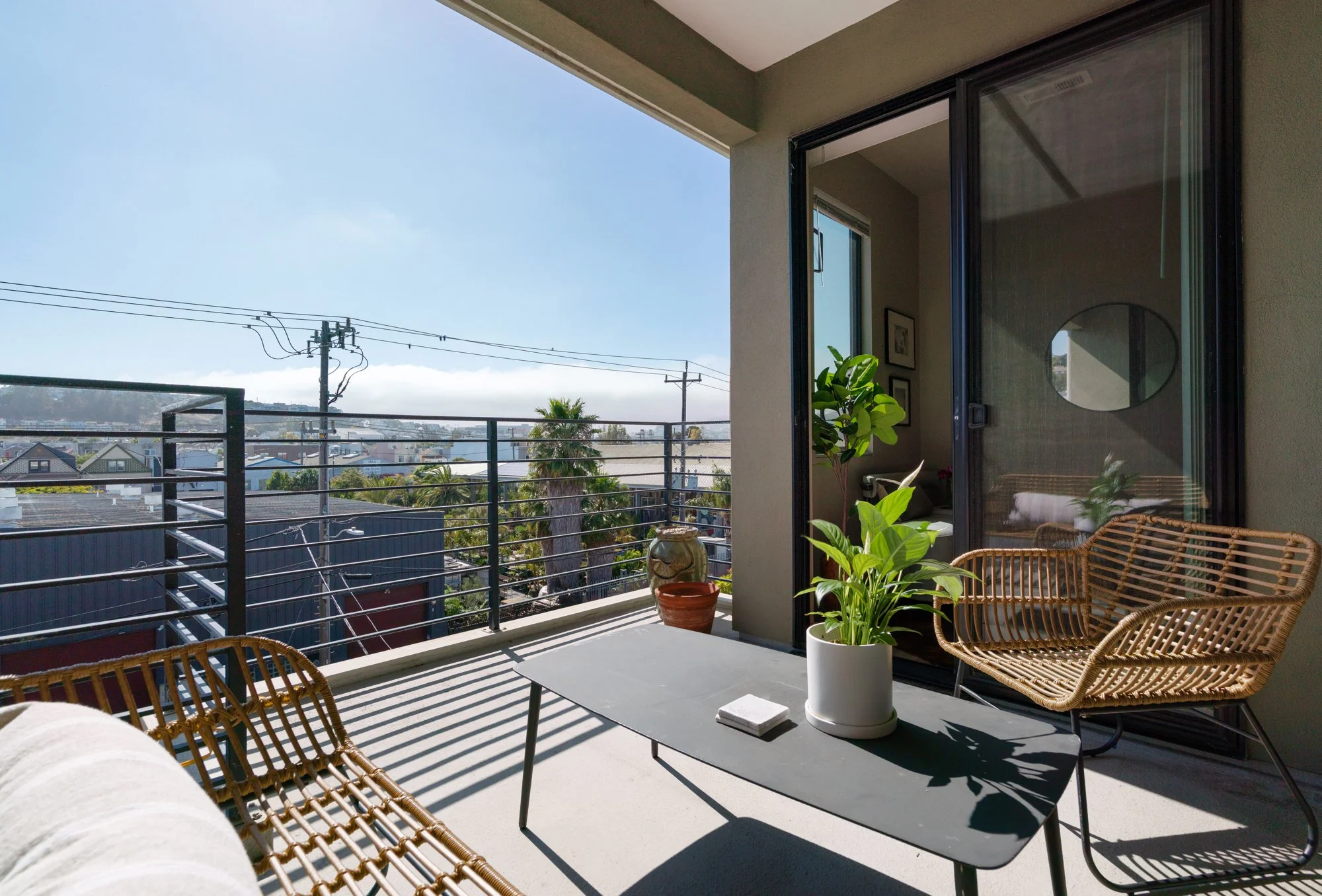 Balcony with wicker chairs, potted plants, and a view of houses, trees, and power lines under a blue sky.
