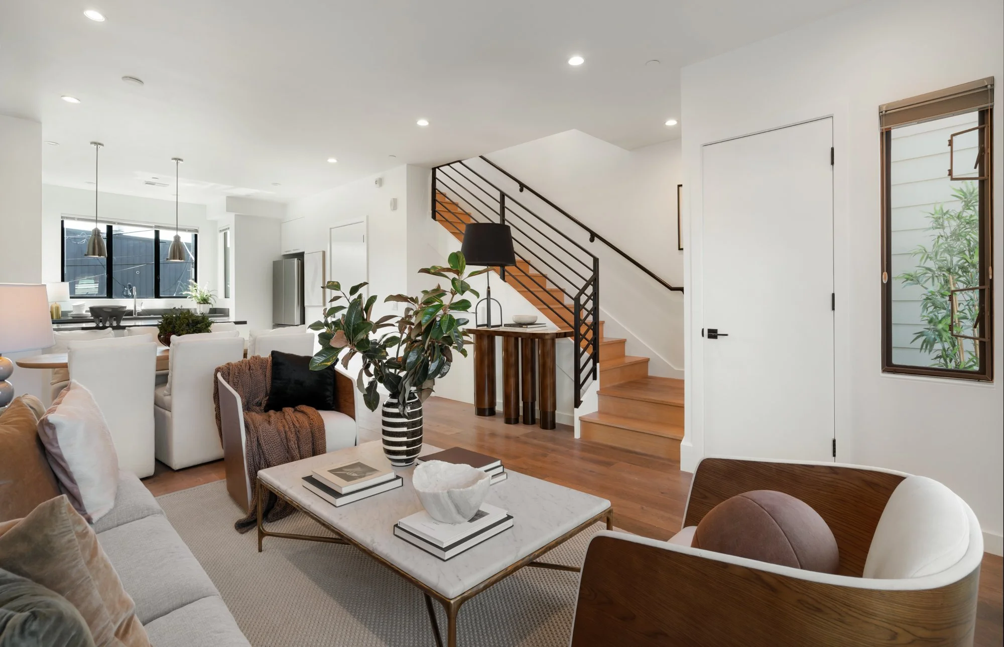Bright living room with white walls and ceiling, wooden flooring, a staircase with black railing, large windows, a sofa with pillows, a coffee table with books and decorative items, and a potted plant.