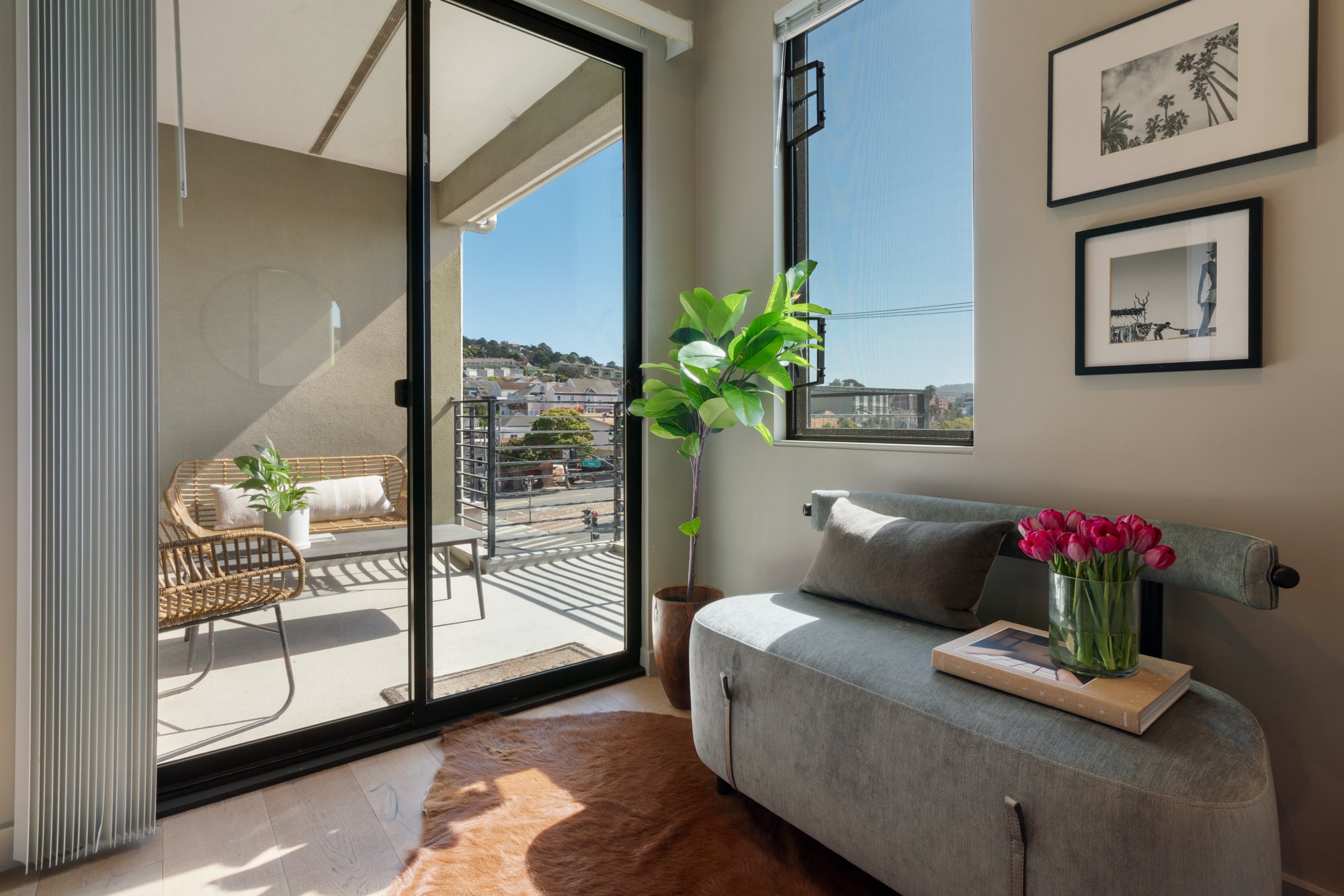 A living room with a sliding glass door leading to a balcony. The room has a gray couch with pillows, a glass vase with pink tulips on a tray, a tall green potted plant, and framed black-and-white photographs on the wall. The balcony features a wicke