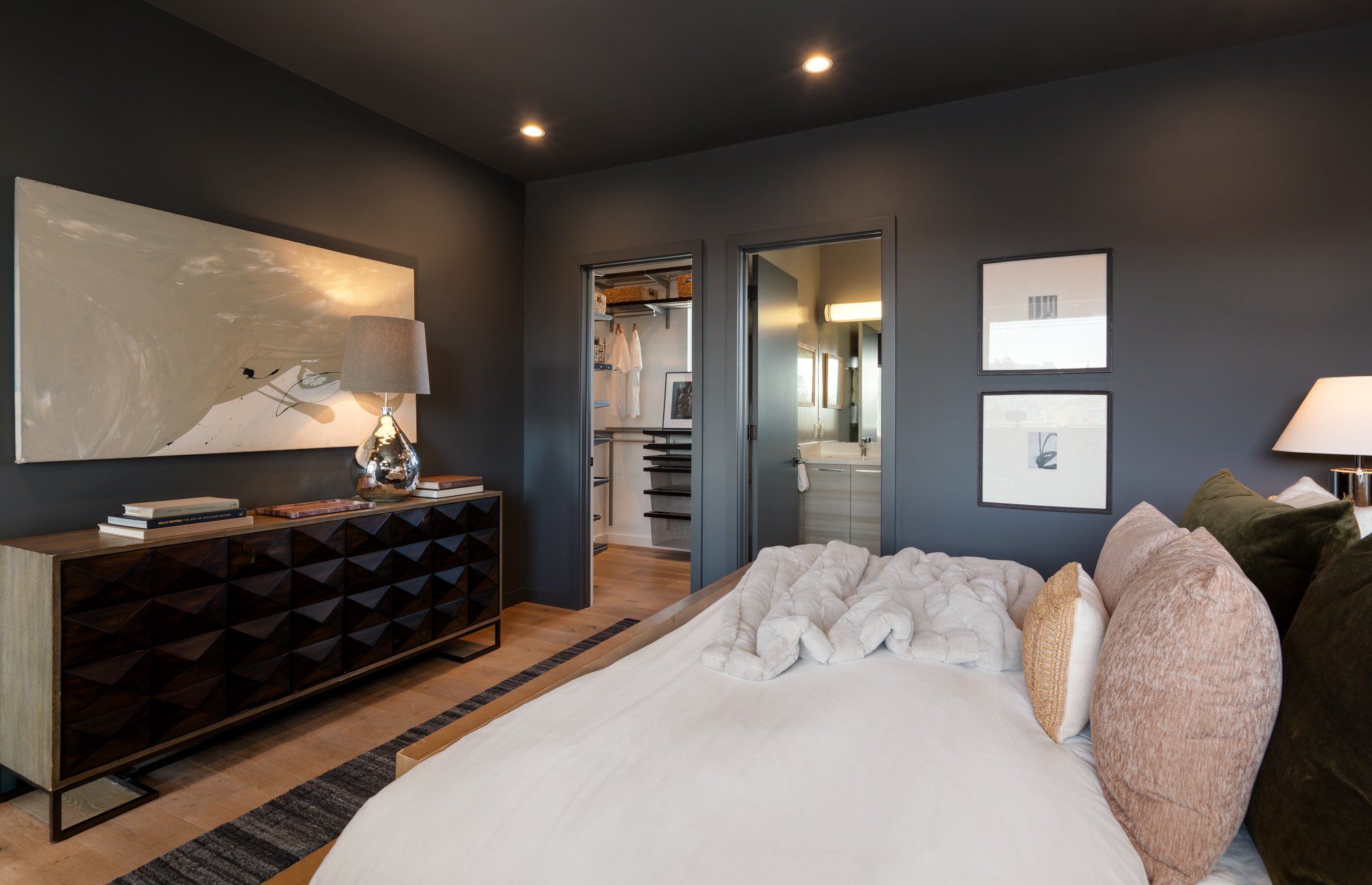 A bedroom with gray walls, a white bed with beige and pink pillows, wooden flooring, a wooden dresser with decorative items, a table lamp, three wall art pieces, two open doors leading to a walk-in closet and a bathroom with a vanity, and recessed ce