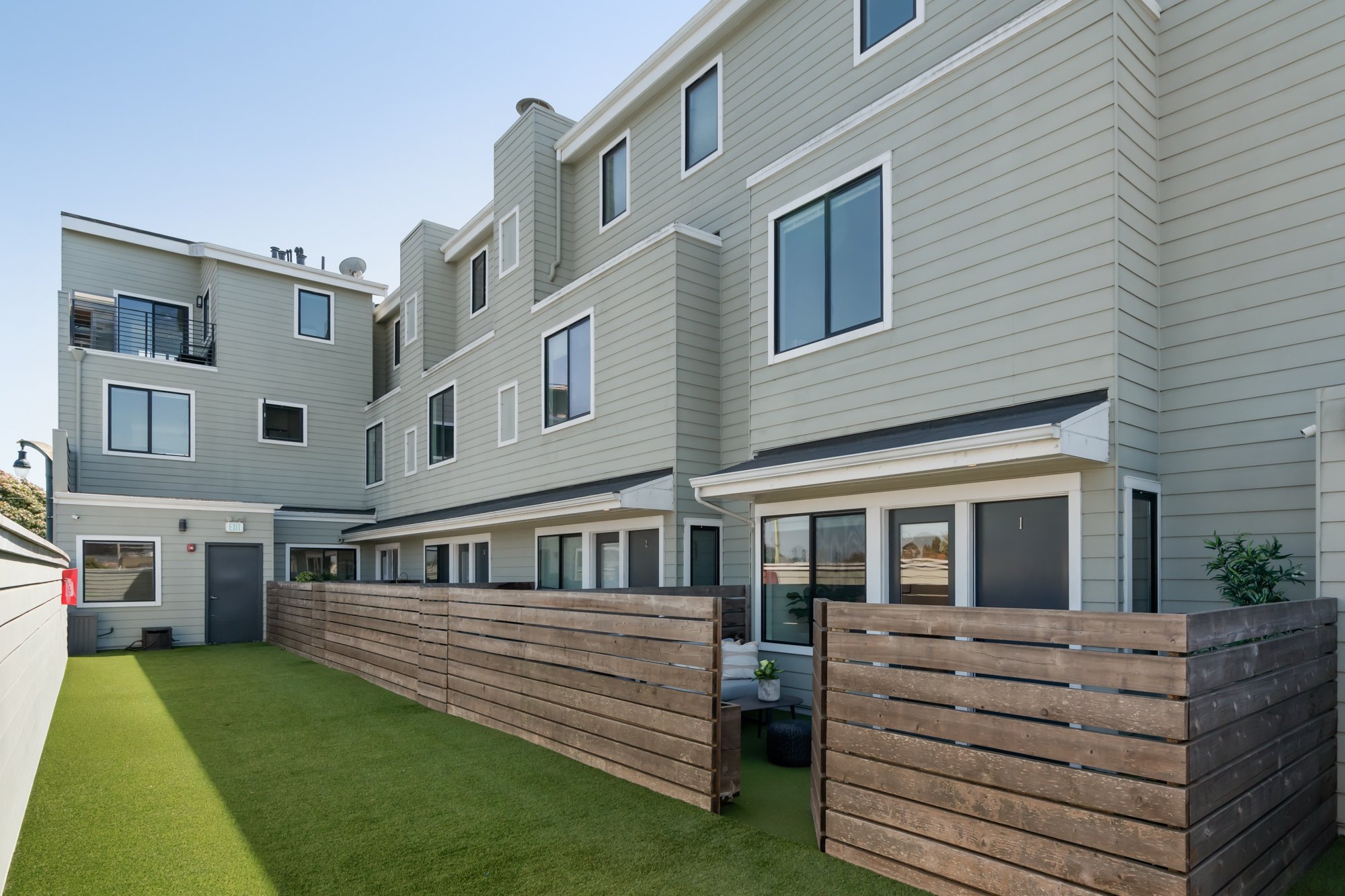 View of a modern multi-story residential building with a small backyard area featuring artificial grass, a wooden privacy fence, and sliding glass doors.