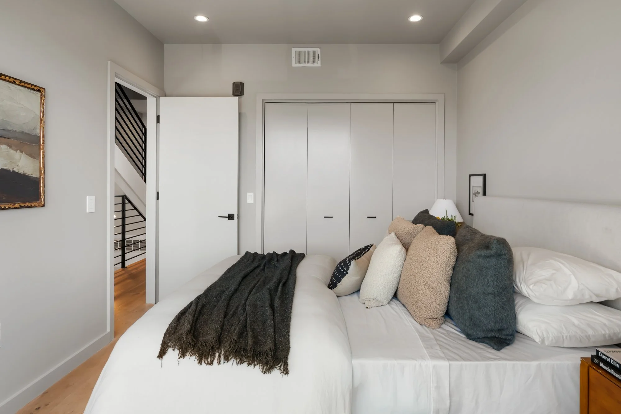 A modern bedroom with white walls, a white bed with multiple pillows, a black blanket, and a mock fur throw. There are two recessed ceiling lights, a painting on the wall, a partially open door showing a staircase, and a closet with sliding doors.
