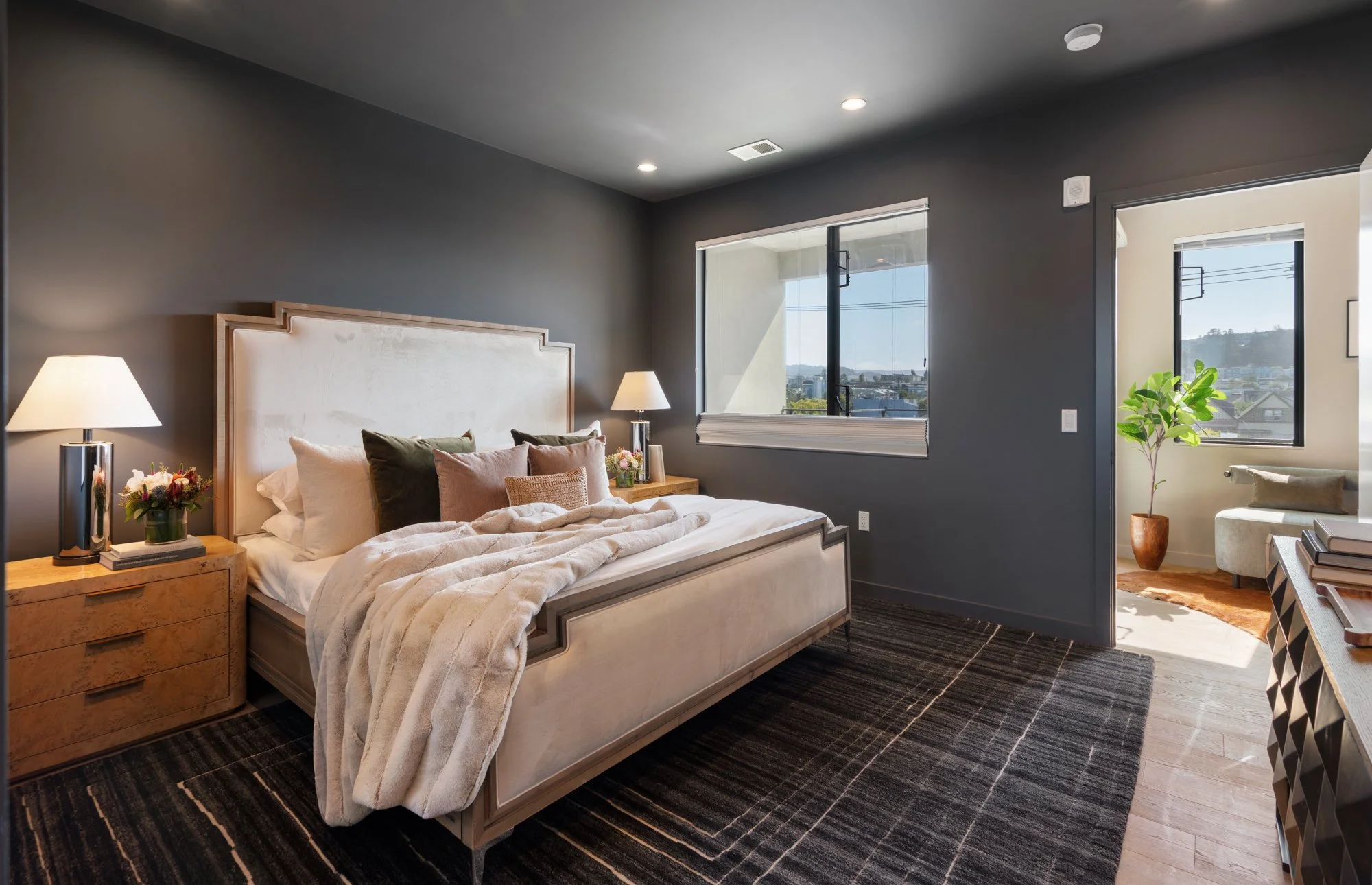Modern bedroom with gray walls, king-sized bed with beige upholstered headboard, white bedding, and green and beige pillows. Nightstands with lamps and flowers. Large window with city view and adjacent room with a window, plant, and chair.