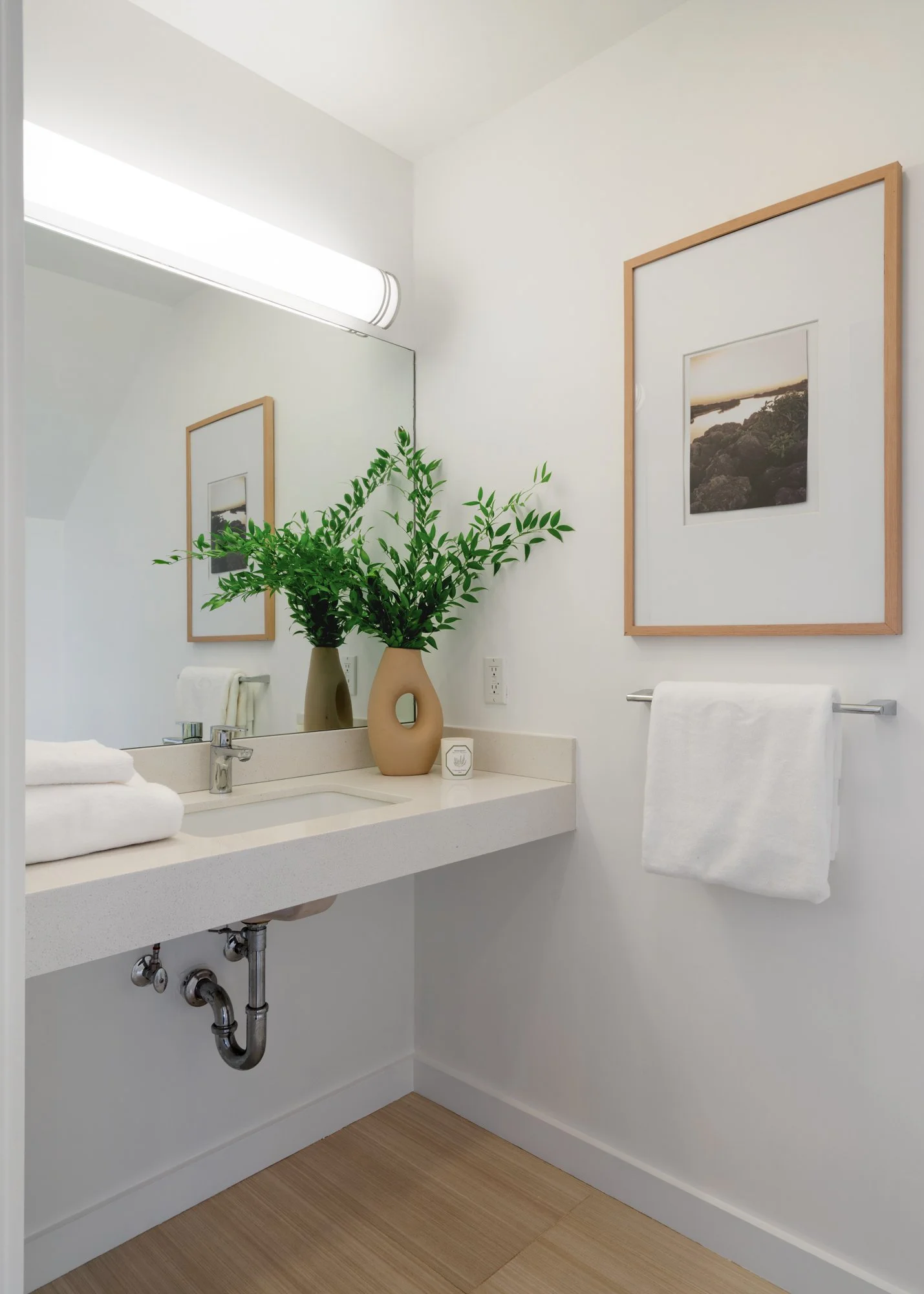 A bathroom vanity with a large mirror, two beige vases with green foliage, a small white candle, and neatly folded white towels. The wall art includes framed landscape photos, and there is a white towel hanging on a silver rod.