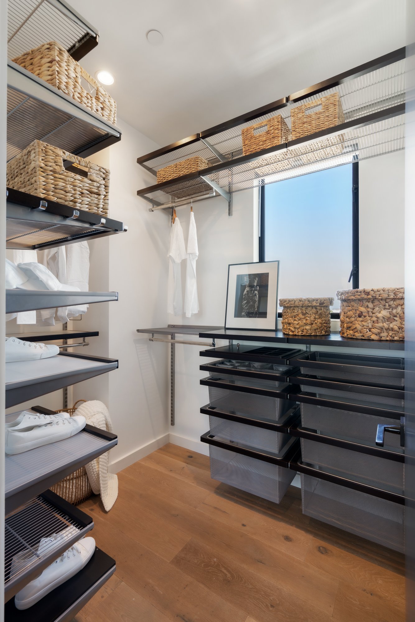 A walk-in closet with white walls, wooden flooring, black wire shelving units filled with woven baskets, white sneakers, and clothes hanging on hooks. There's a framed photo on a black counter, shelves with additional baskets, and a window with a bla