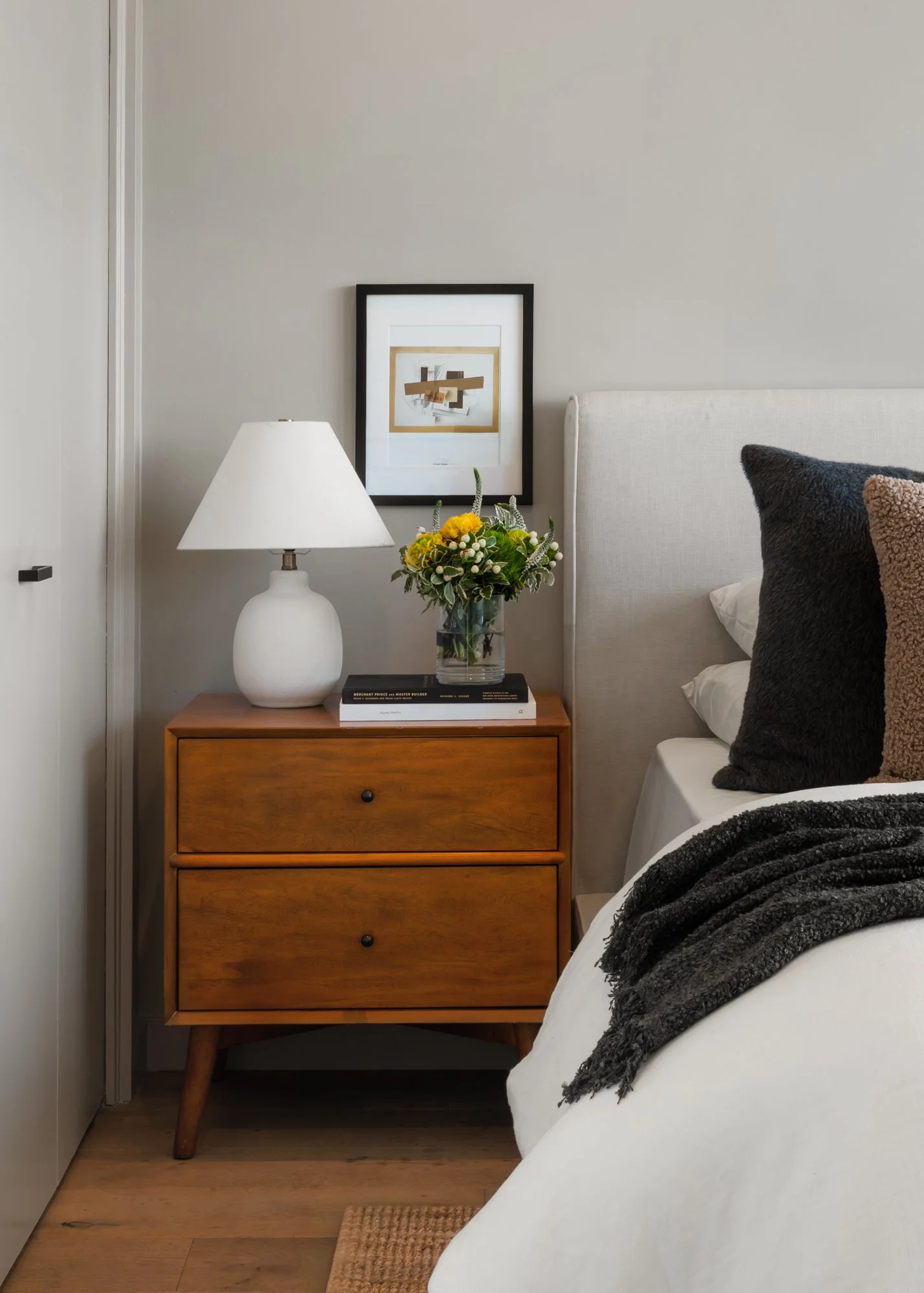 A bedroom corner with a wooden nightstand holding a white table lamp, a glass vase with yellow flowers, and two books. Above the nightstand, a framed abstract artwork hangs on a light gray wall. Part of a white bed with black and brown pillows and a 