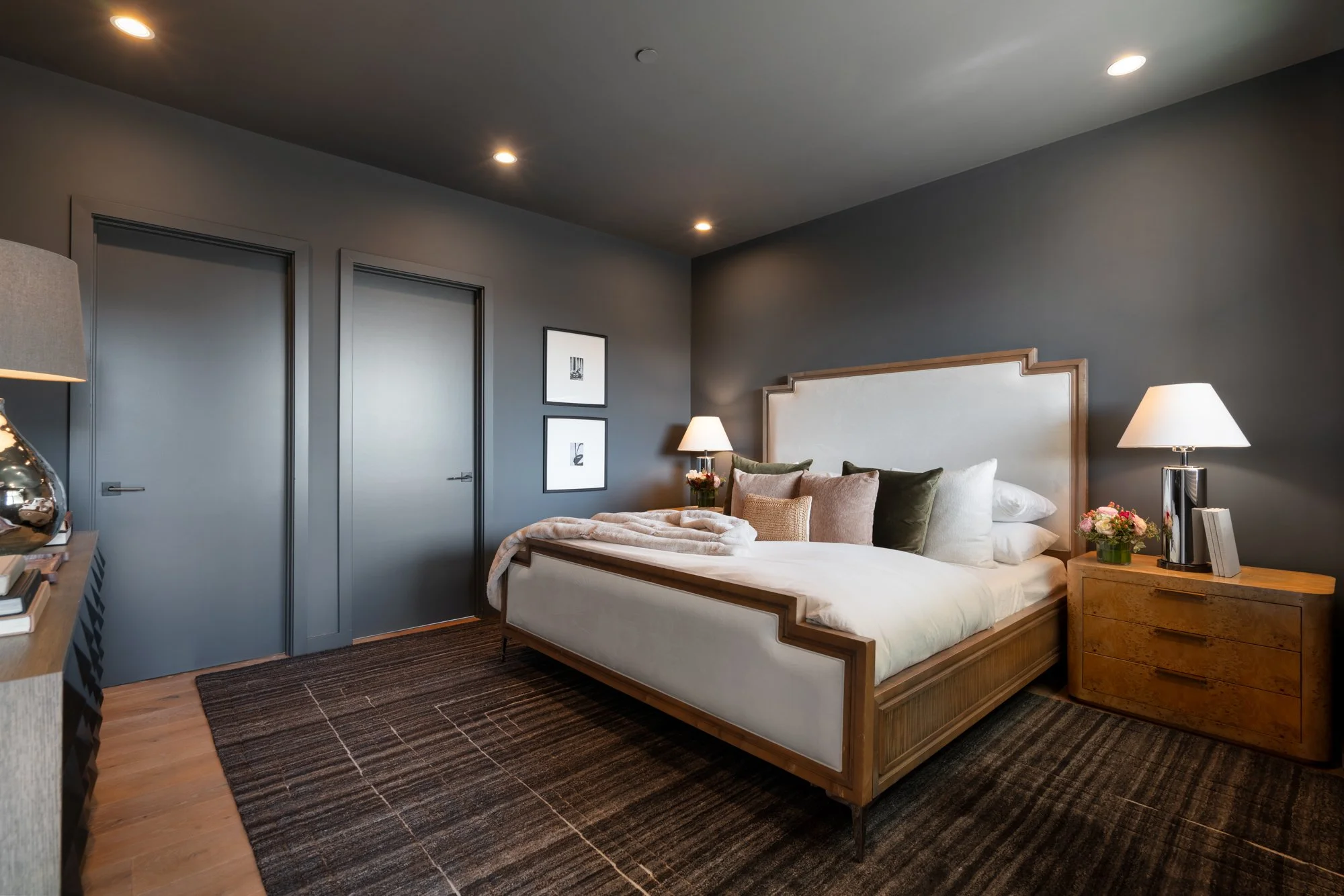 A modern bedroom with a wooden bed frame, white upholstered headboard, beige and green throw pillows, nightstands with lamps, and framed art on gray walls.