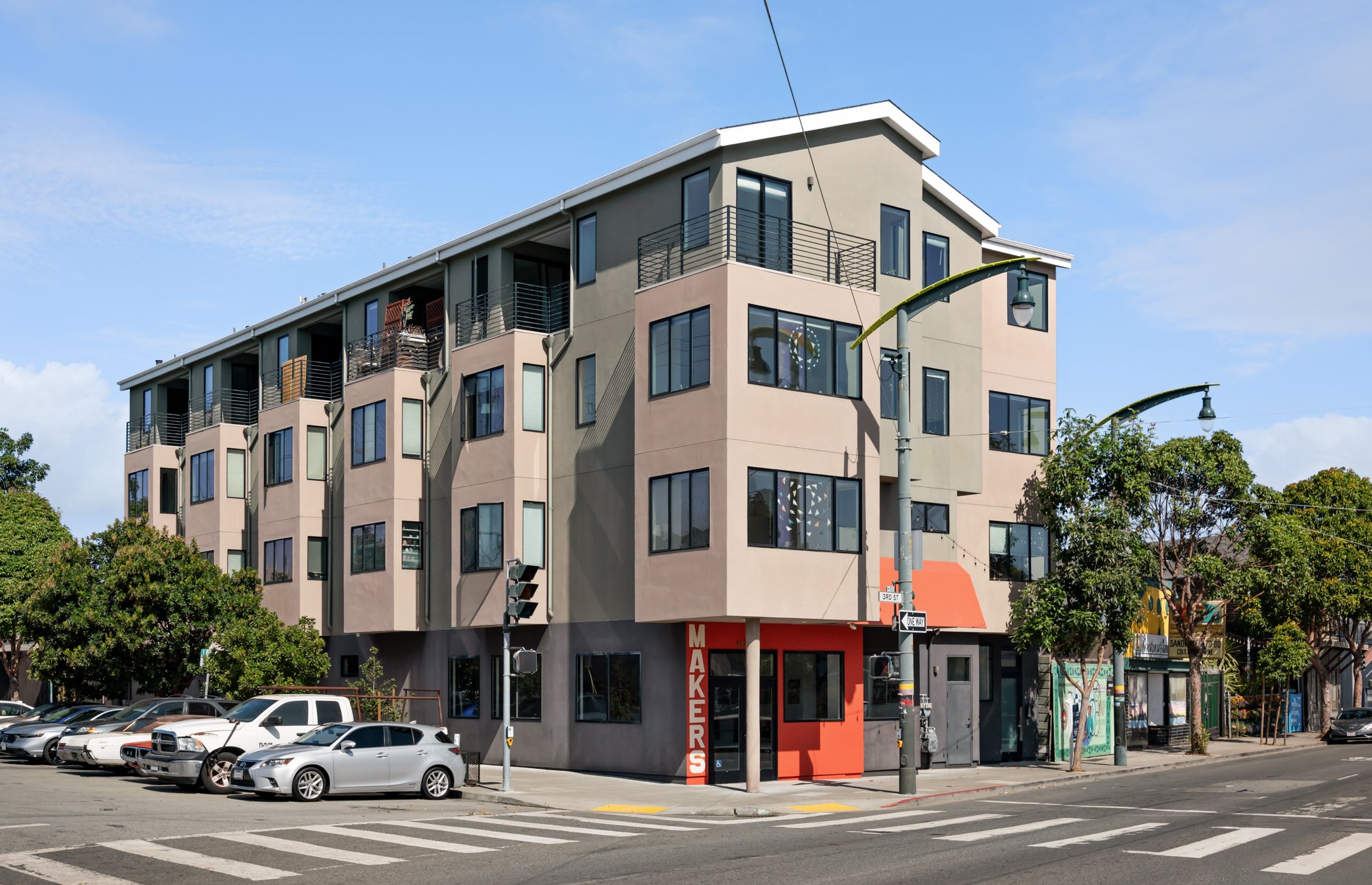 A modern multi-story apartment building with multiple balconies, large windows, and a corner storefront with a red sign that reads "MAKEERS." The scene includes parked cars, street lamps, trees, and a crosswalk on a clear day.