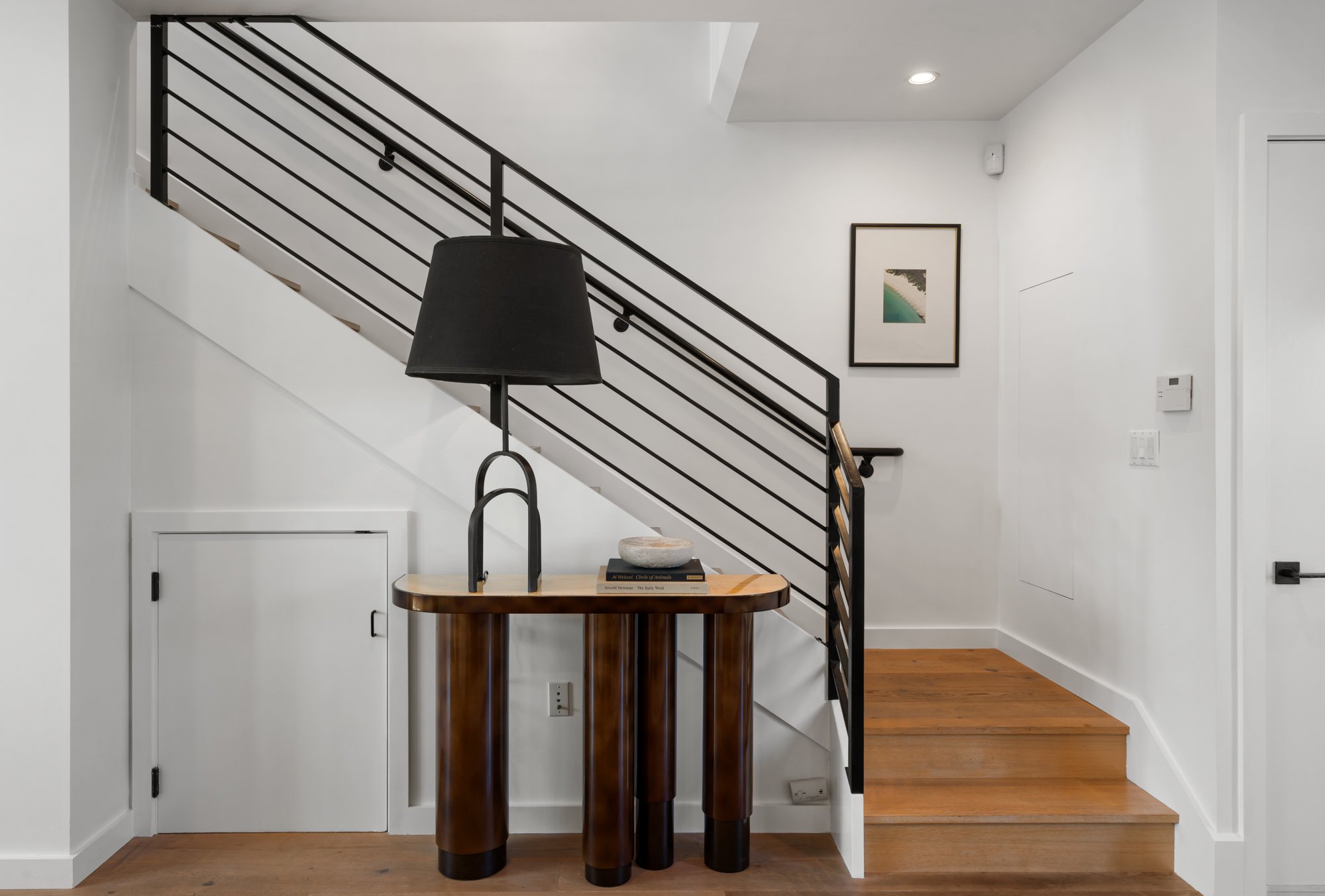 Interior view of a modern staircase with black metal railings, wooden steps, and a small wooden table with a black lamp, a book, and a decorative bowl on it. There is a framed picture on the wall and various control panels.