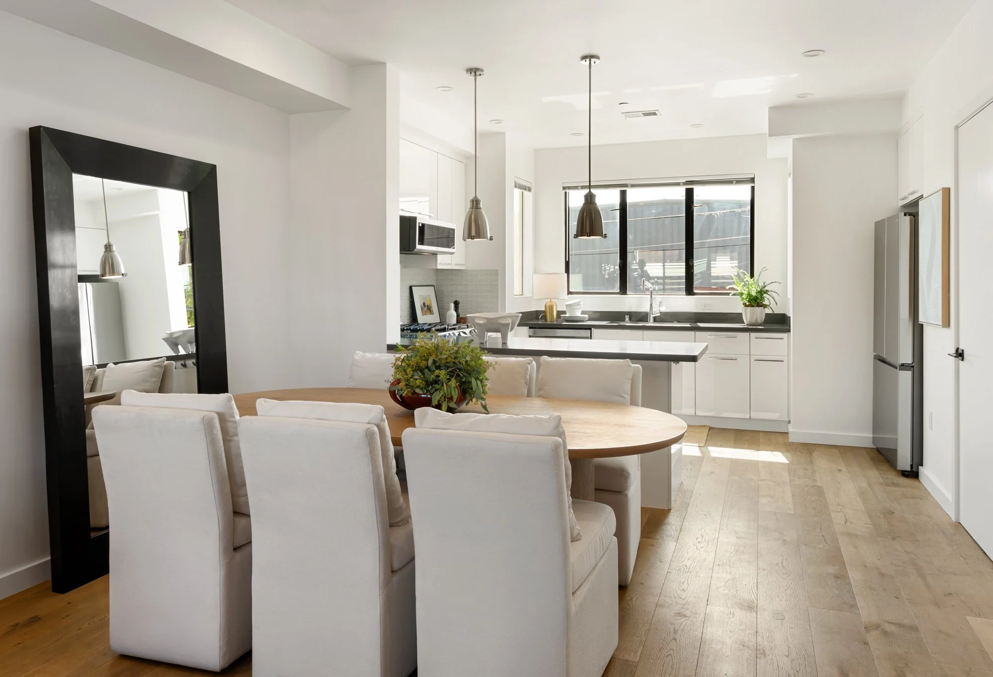 Modern white kitchen with large windows, black accents, and a dining area with a round wooden table and white chairs.
