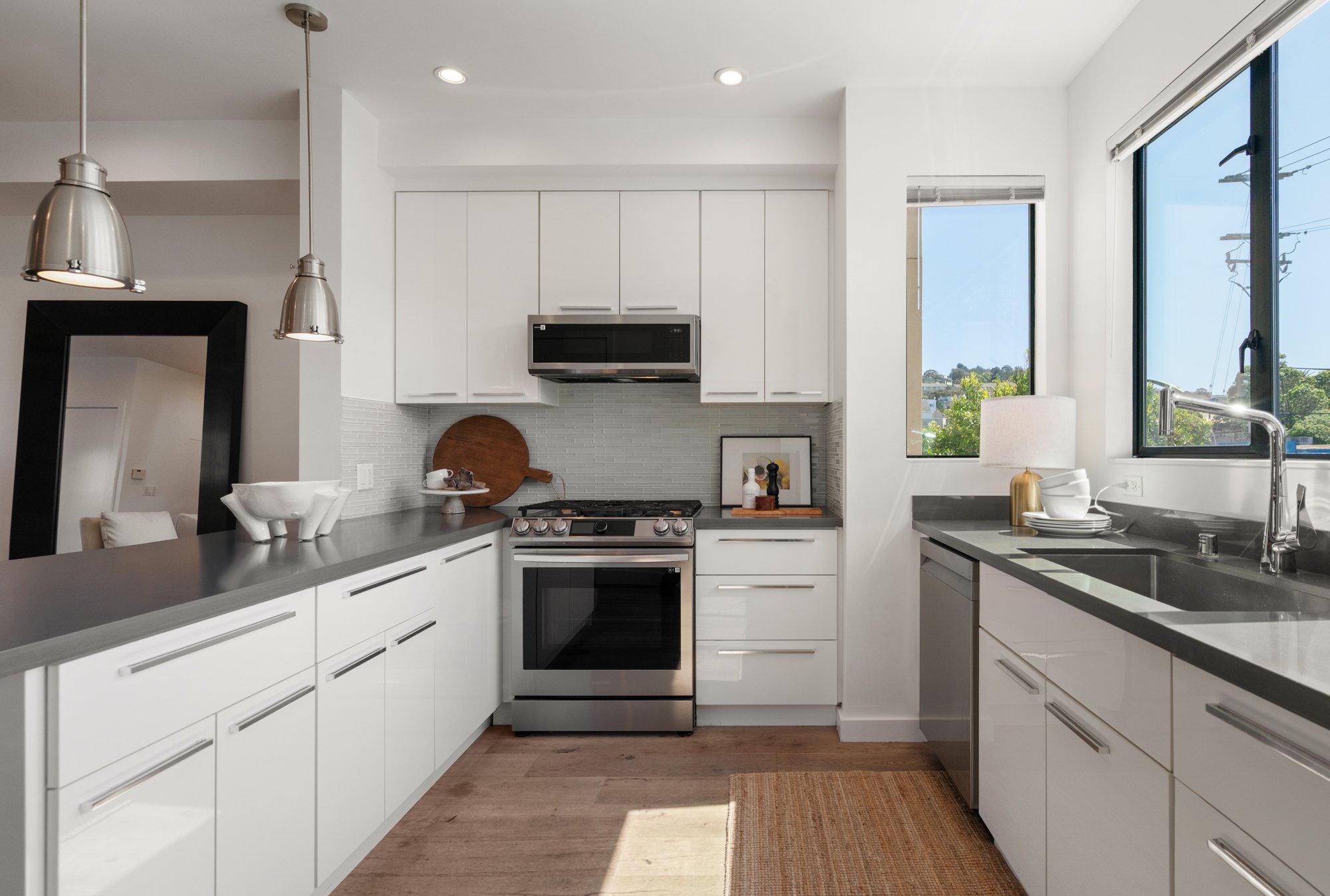 Modern kitchen with white cabinets, stainless steel appliances, large windows, and wooden flooring.