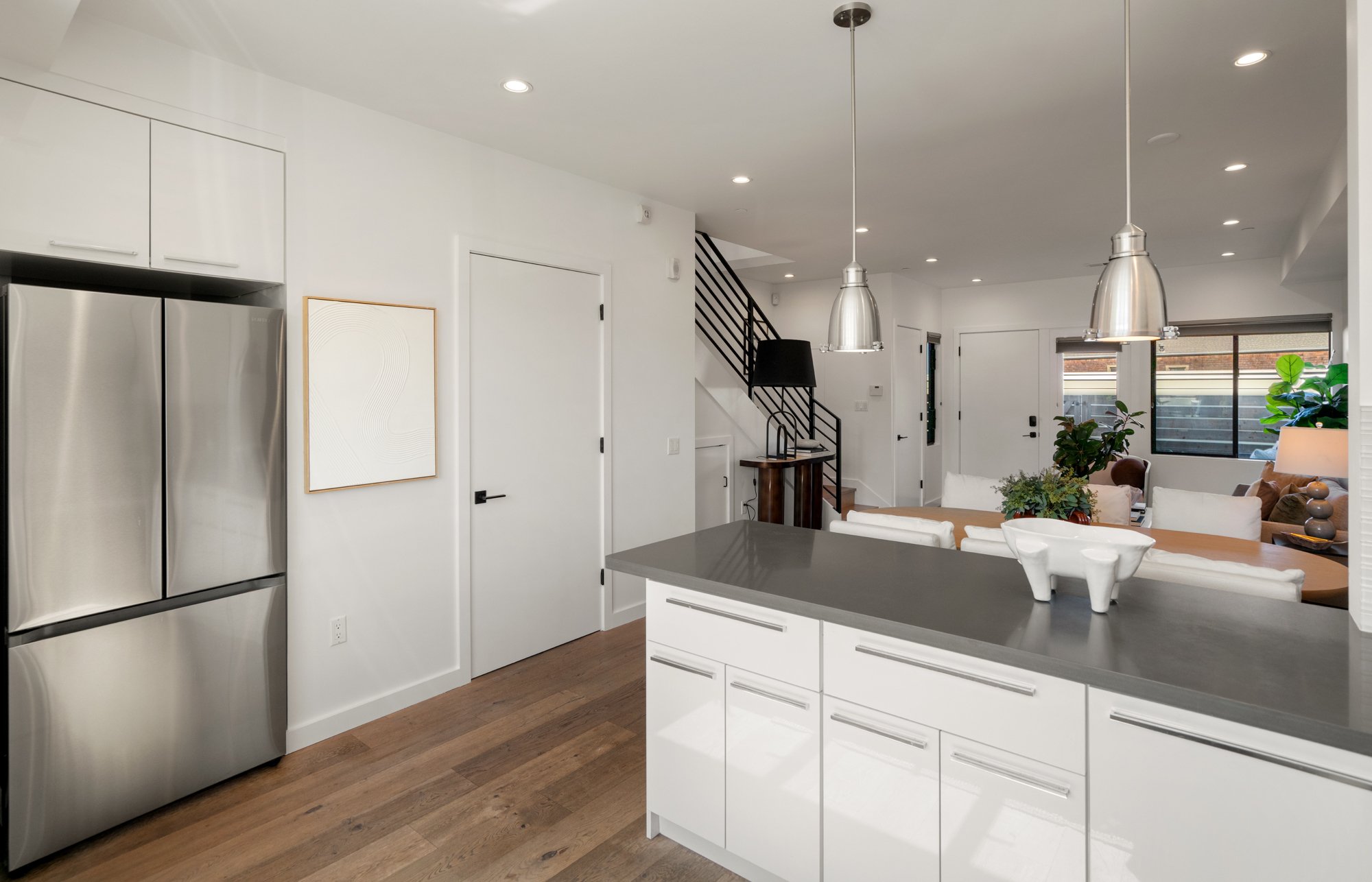 A modern open-concept kitchen and living space with white cabinetry, stainless steel refrigerator, wood flooring, and minimalist decor. There are two metallic pendant lights hanging over a kitchen island, and a staircase leading to the upper floor.
