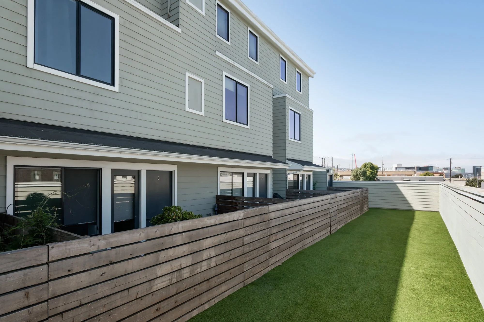 A multi-story residential building with beige siding, black-framed windows, and a backyard with a wooden fence, small green plants, and a well-manicured lawn under a clear blue sky.
