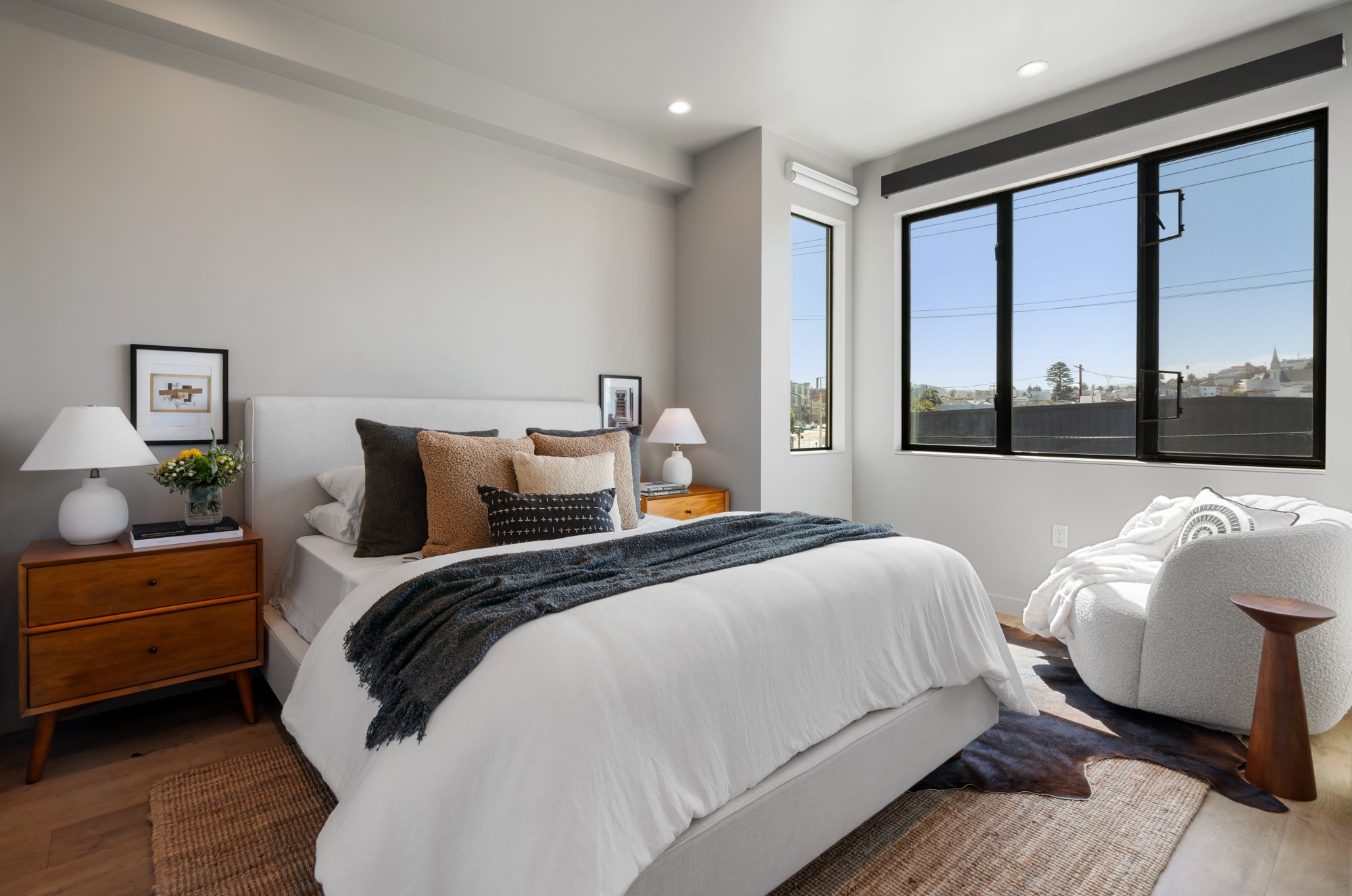 A modern bedroom with a large bed featuring white linens and an assortment of black, tan, and cream pillows. Wooden nightstands with white lamps and decorative framed artwork. Large windows with black frames provide a view of a cityscape with houses,
