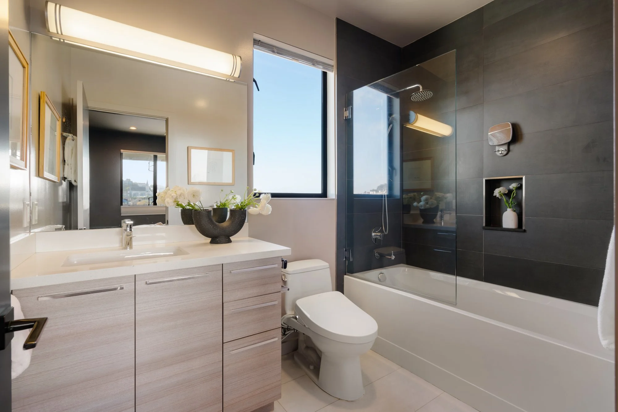 Modern bathroom with light wood vanity, white sink, large mirror, vase with white flowers, dark tile shower with glass door, window with city view, toilet, and bathtub.