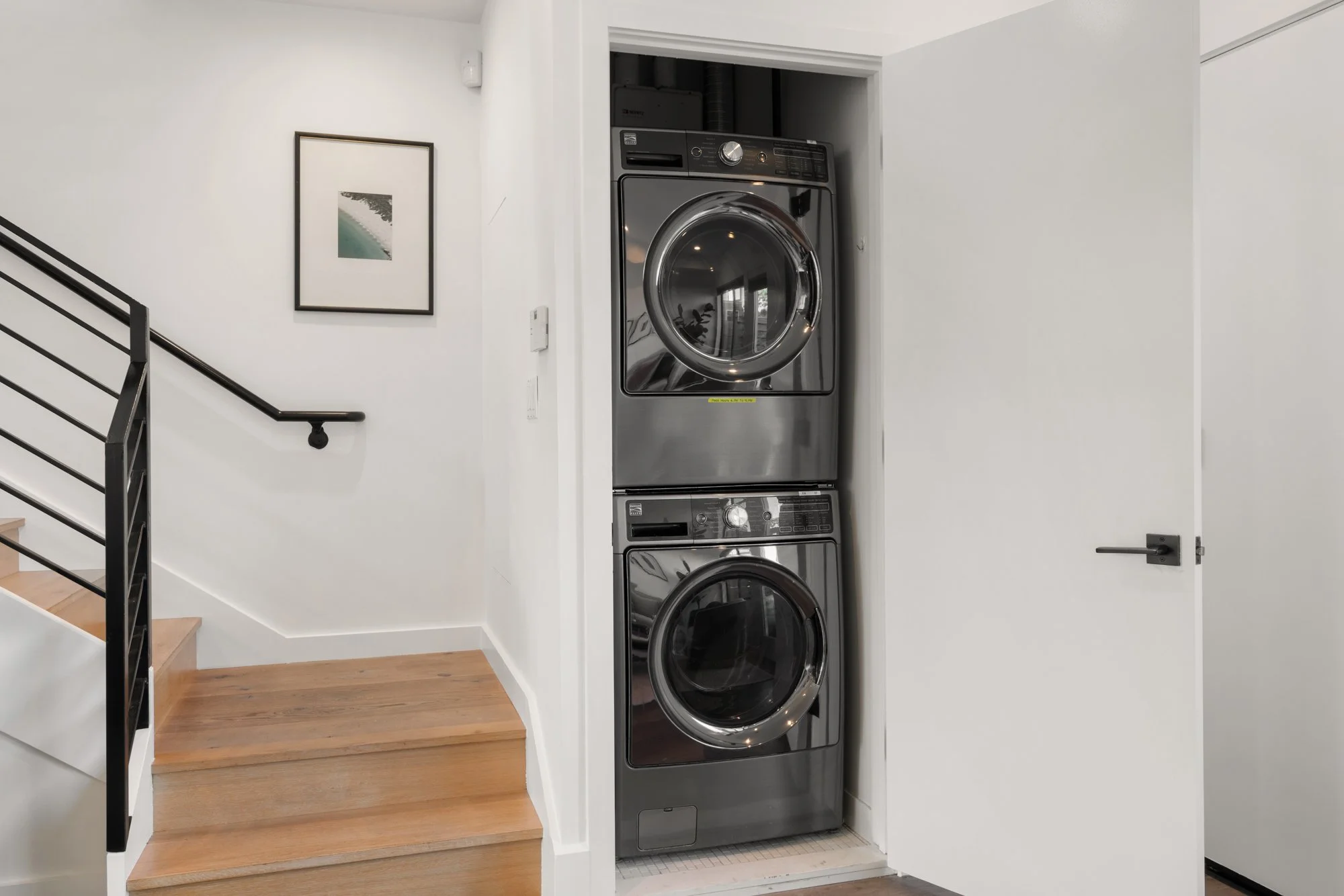 Stacked metallic front-loading washer and dryer in a small, recessed laundry space next to a white. wall and a door.