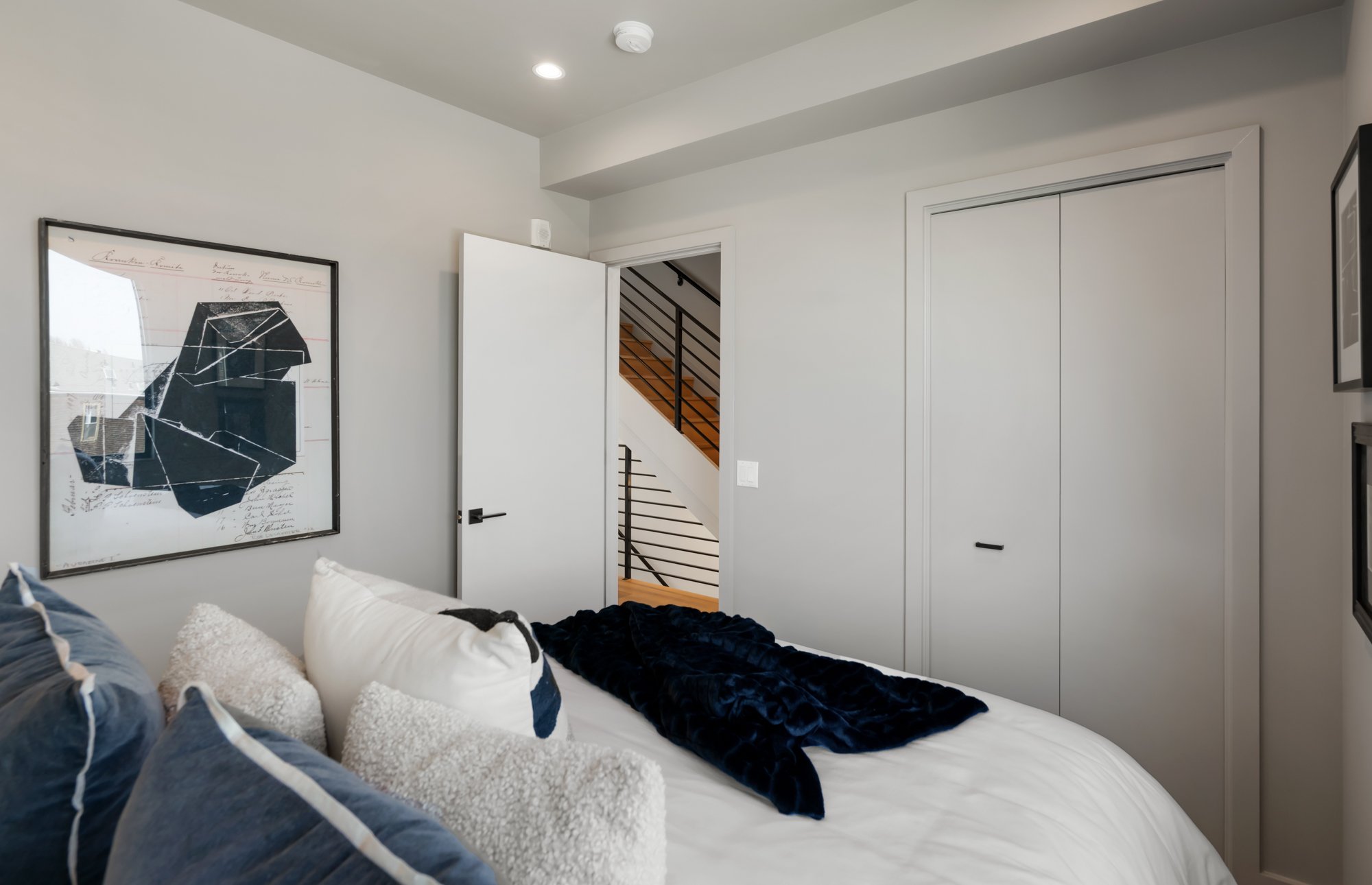 A bedroom with a bed that has white and navy blue pillows and a black blanket, a framed abstract art piece on the wall, a white door open revealing a staircase with black railing, and a closet with white sliding doors.