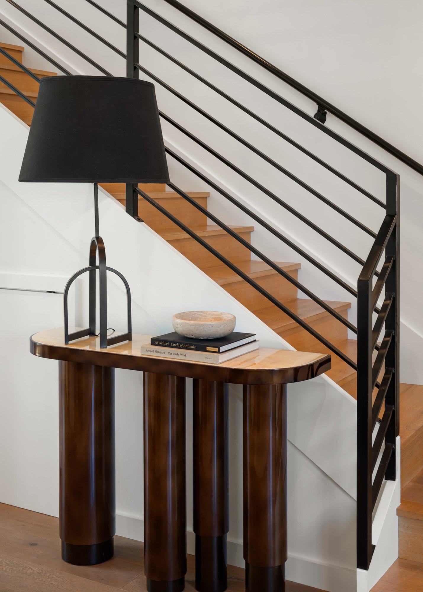 Interior view of a modern staircase with black metal railing, wooden stairs, and a white wall. In the foreground, there is a black lampshade on a tall black lamp and a wooden table with books and a decorative stone bowl.