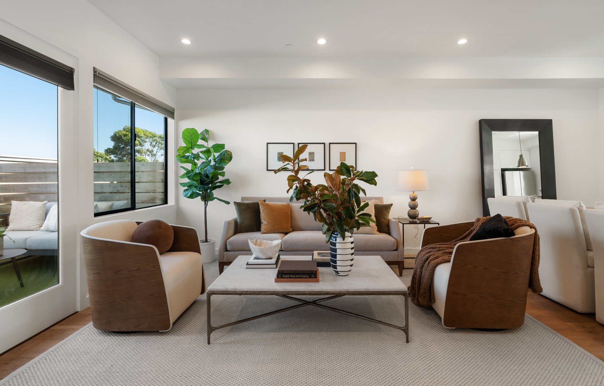 A modern living room with white walls and a ceiling with recessed lighting. Large windows with blinds allow natural light, and there are green plants and framed artwork on the wall. The room has a beige sofa with pillows, a coffee table with books an