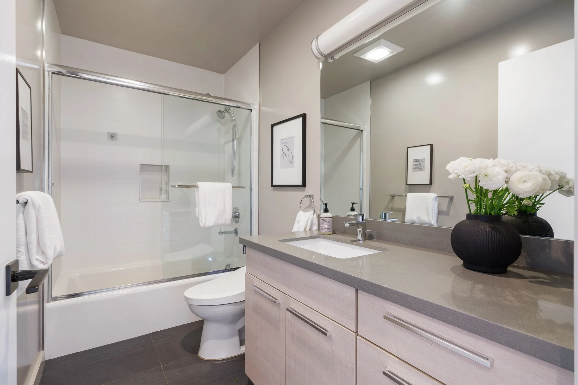 Modern bathroom with a glass-enclosed shower, white towels, a toilet, a beige vanity with a sink, a large mirror, a black vase with white flowers, and framed artwork on the walls.