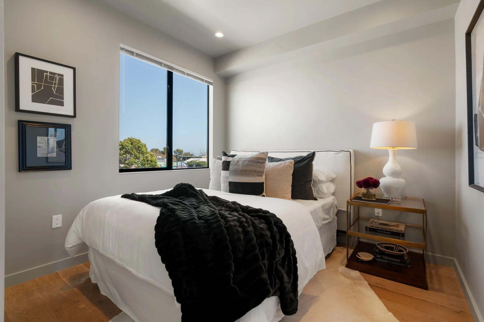 A bedroom with a bed covered in white bedding and a black blanket, multiple pillows, a side table with a white lamp, a small bouquet of flowers, and books, with artwork on the wall and a window showing a view of trees and rooftops outside.