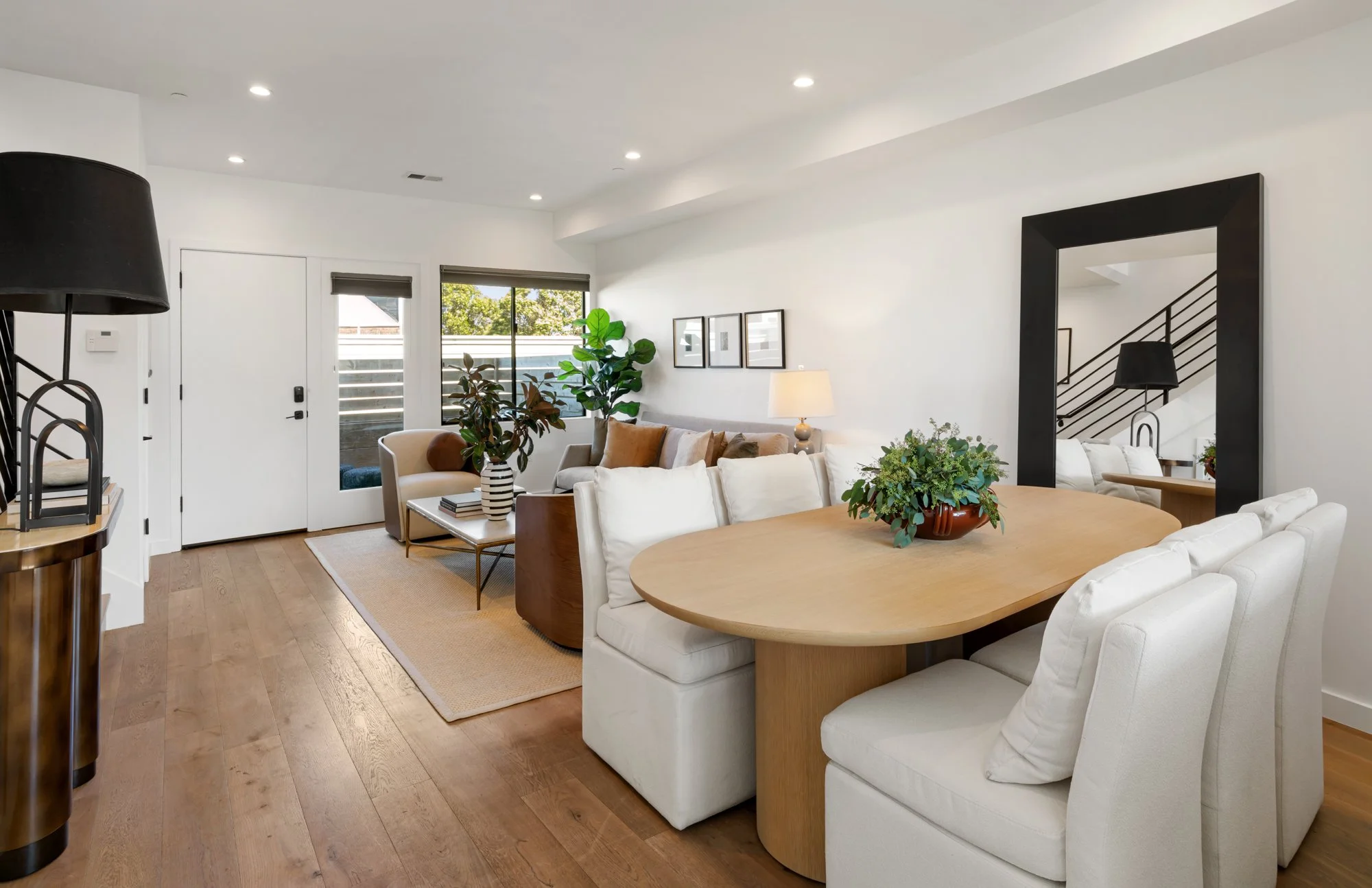 Bright, modern living room with white walls and hardwood floors, featuring a large beige sofa with brown pillows, a wooden dining table with white upholstered chairs, a black-framed mirror, plants, and framed artwork on the wall.