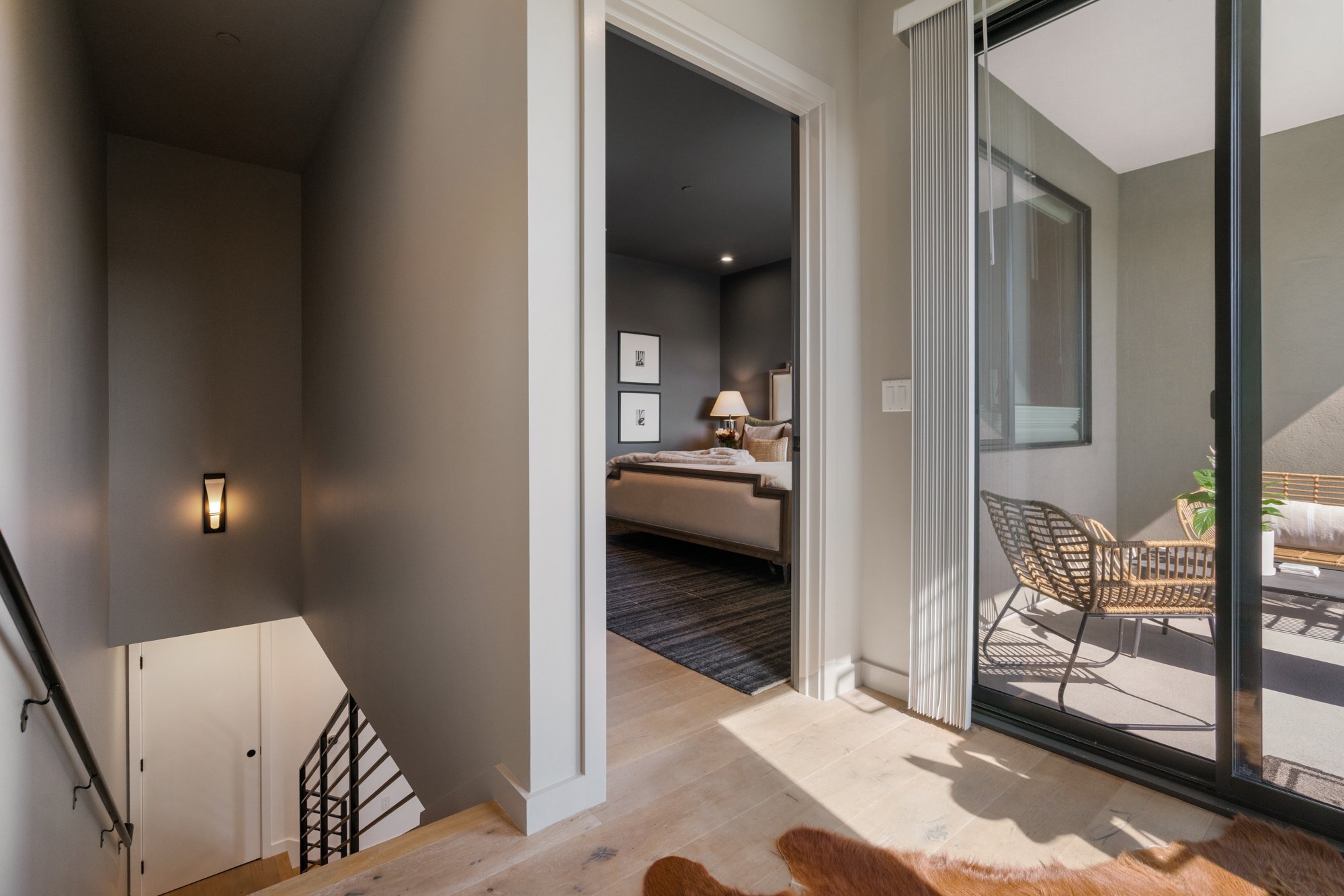 View from top of stairs showing a hallway leading into a bedroom with gray walls, hallway with natural light, balcony with glass sliding door, outdoor table with chairs, small indoor lamp on bedside table, framed pictures on bedroom wall, hardwood fl