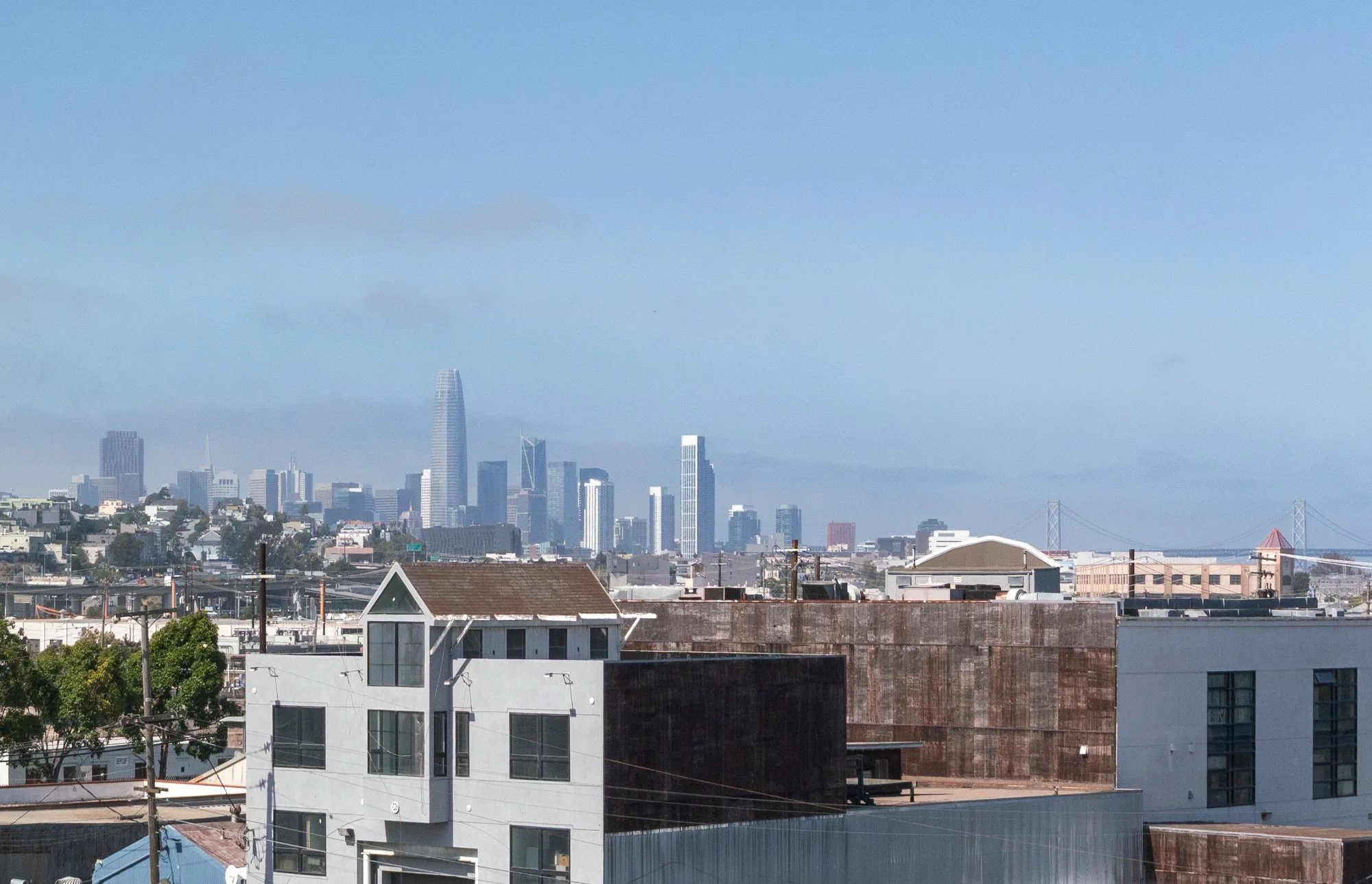 View of a city skyline with tall skyscrapers in the distance, residential houses and rooftops in the foreground, and a bridge partially visible on the right, under a blue sky.