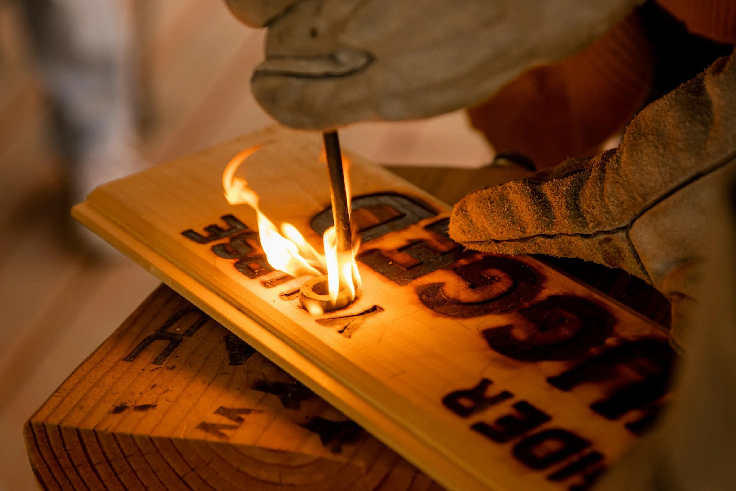 Close-up of a wooden block with the word 'LOVE' burned into it, on which there is a small flame. The background shows some rough-textured material.