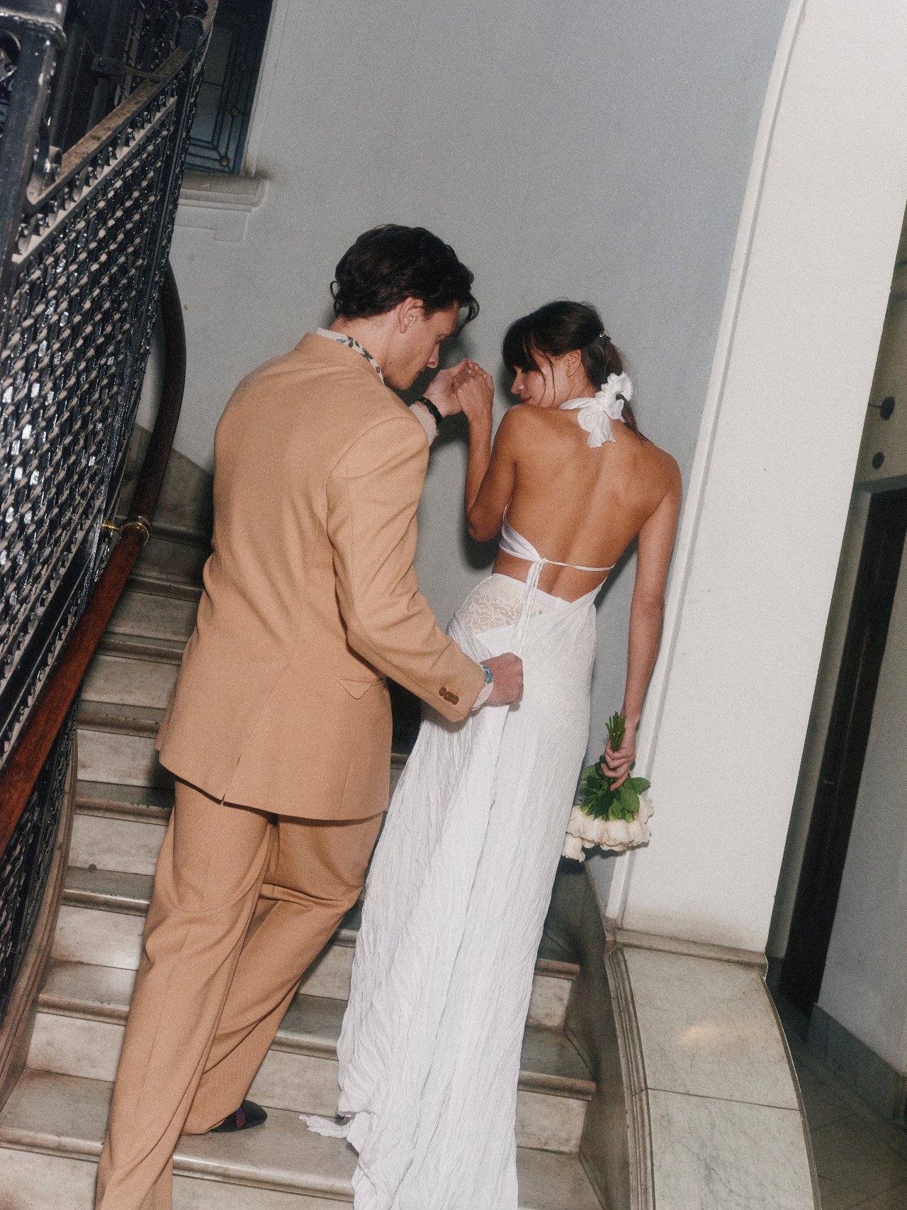 A man in a tan suit holding the hand of a woman in a white dress who is holding a bouquet of flowers, as they walk up a staircase.