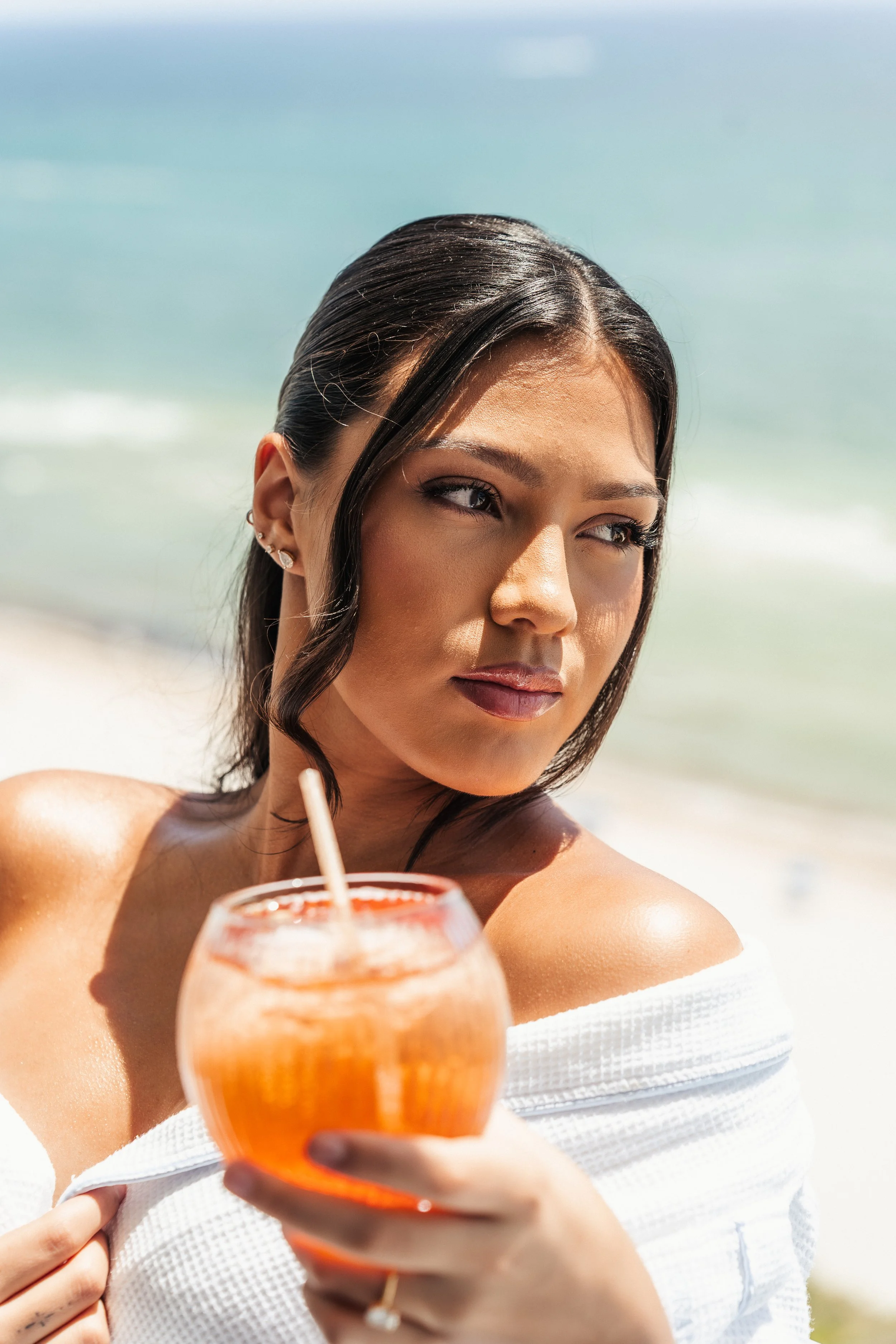 Close-up of a young woman with dark hair and earrings, holding a drink with a straw at the beach, with ocean and sky in the background.