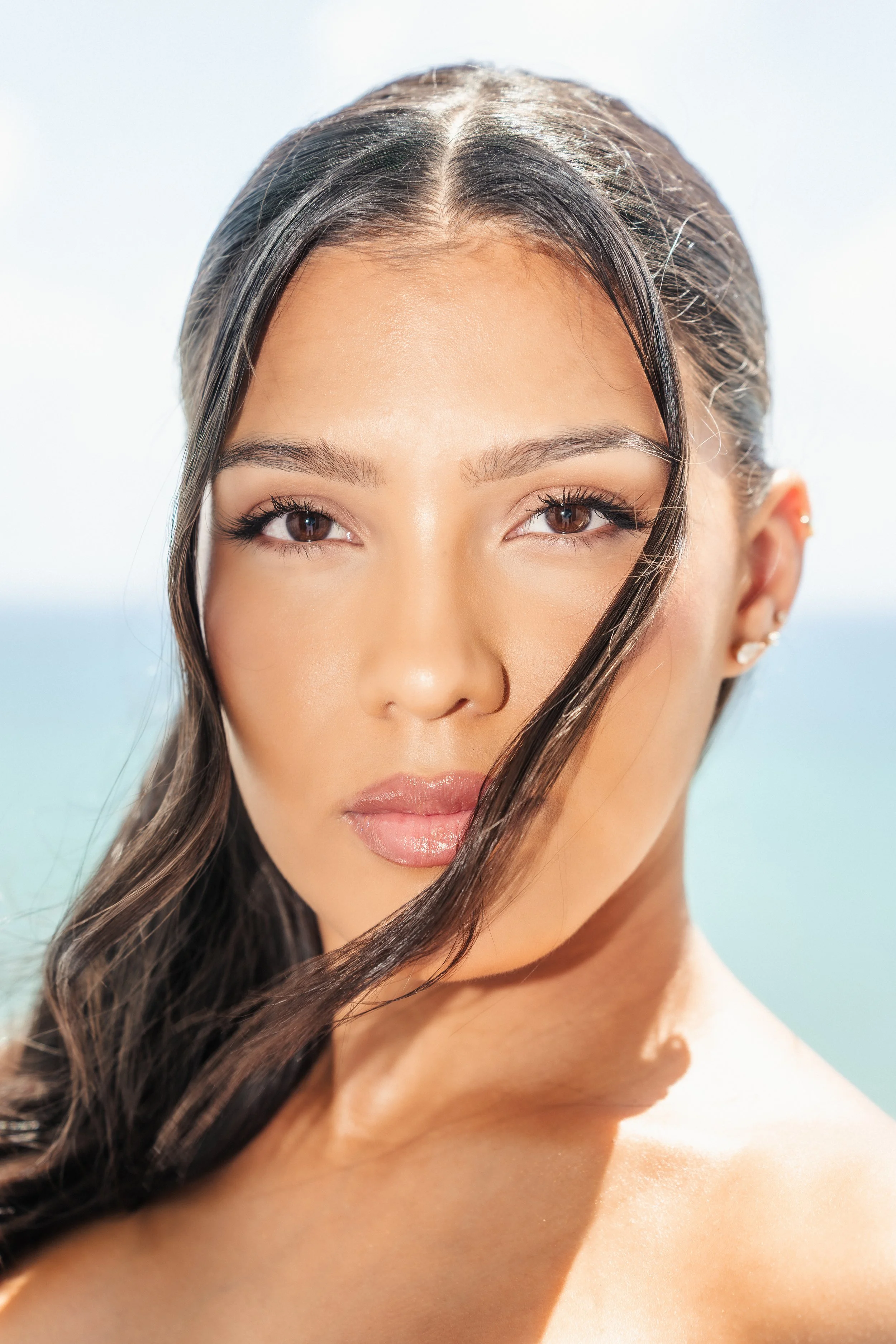Close-up of a woman's face with brown eyes, long dark hair, and smooth skin, outdoors with a bright sky background.
